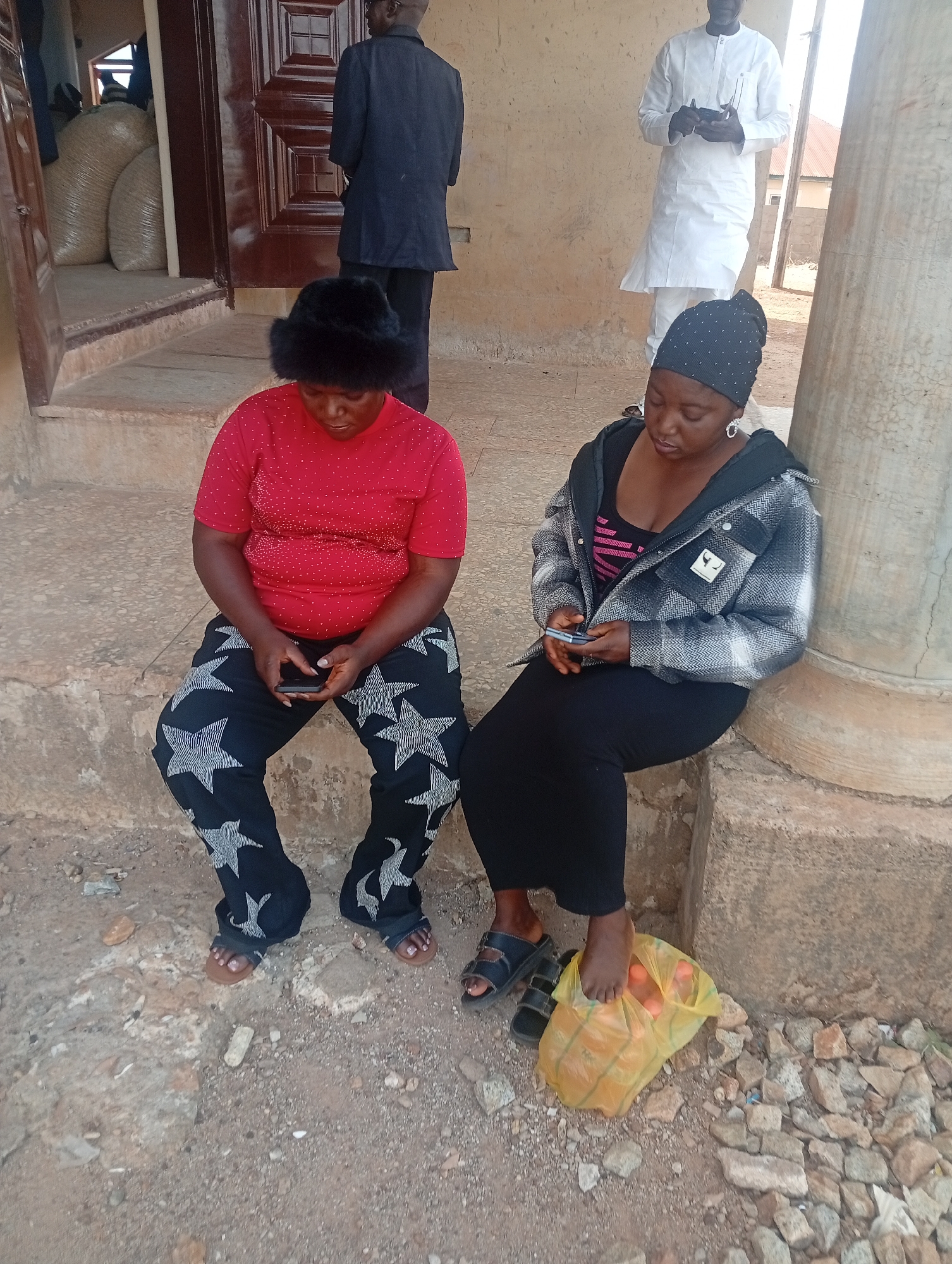 This image depicts a casual outdoor scene in Ladura Foron, Nigeria, captured during what appears to be bright daylight. The foreground is dominated by two women seated on a low concrete ledge or step.
The woman on the left is wearing a red short-sleeved top adorned with small white embellishments, and black pants featuring large, prominent white star patterns. She has a black furry hat on her head and is barefoot, with light brown sandals on the ground beside her feet. She is holding a mobile phone in both hands, looking down intently at its screen.
Beside her, to the right, sits another woman. She is dressed in a black and white plaid jacket over a dark top, a long black skirt, and a black beanie cap embellished with small studs. She is also focused on a mobile phone held in her hands. Her bare right foot rests on a yellow translucent plastic bag, which contains several oranges or similar round fruits. She wears black sandals. A substantial concrete pillar is visible to her right.
In the background, two men are standing. One man, seen from behind, wears a dark suit jacket and trousers, positioned near a large, ornate wooden door. Through the open doorway, several large, light-colored sacks, possibly filled with grain or other goods, are visible. Further to the right, a second man stands in a traditional long white garment, holding a small object in his hands, likely another mobile phone.
The ground surface in the immediate vicinity is a mixture of dirt and small rocks or gravel. The architecture includes light-colored, possibly painted concrete walls and steps. The overall impression is one of people waiting or resting in an open-air or semi-covered area, engaging with their personal devices. No discernible text is visible within the image.