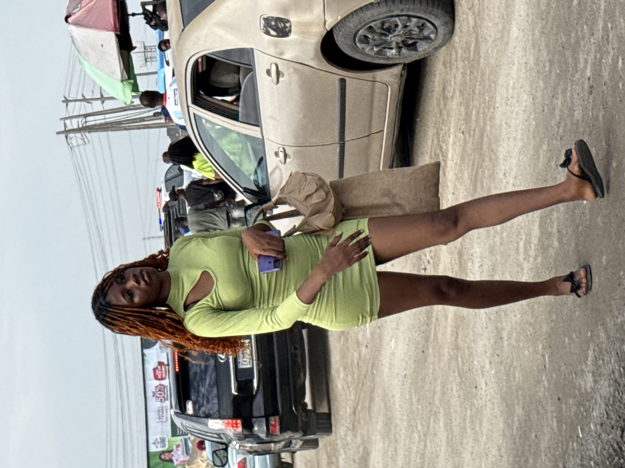 A woman with reddish-brown hair is standing on a street in Lekki, Nigeria. She is wearing a light green, long-sleeved mini dress and black sandals. She is holding a blue cell phone in her left hand. A light brown tote bag hangs off her shoulder. Behind her are cars, including a dark-colored SUV. In the background, there are utility poles, advertising signs, and other pedestrians. A car is partially visible at the top of the frame, and a street vendor with an umbrella is also partially visible in the background. The ground is a concrete or paved surface.