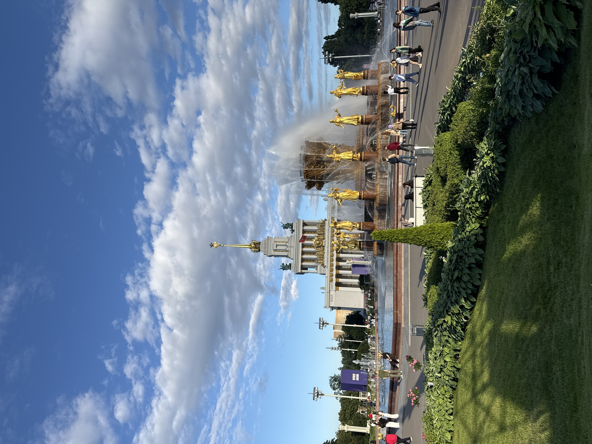 The image captures a scene at the VDNKh (Exhibition of Achievements of National Economy) park in Moscow, Russia. In the foreground, a manicured lawn with dappled sunlight creates a welcoming atmosphere. Behind the lawn, a large fountain with golden statues of women is in full operation, with water spraying high into the air. The iconic Friendship of Peoples Fountain, a central attraction at VDNKh, is prominently featured. In the background, a grand pavilion, likely one of the Soviet-era exhibition halls, stands tall with classical columns and intricate golden decorations. The sky above is a vibrant blue, dotted with fluffy white clouds, indicating a pleasant day. Several people are walking around the fountain area and on the pathways, enjoying the park. Some are dressed casually, suggesting it's a popular spot for recreation and sightseeing. The overall impression is one of a grand, historic public space bustling with activity on a clear day.