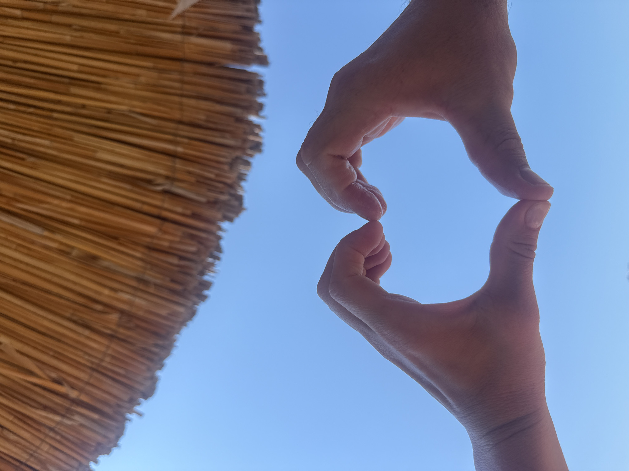 The image captures a close-up of two hands forming a heart shape against a clear blue sky. Above the hands, the thatched roof of a beach umbrella is visible, its dry, straw-like material casting a rustic texture. The hands, positioned centrally in the frame, are illuminated from behind, with their outlines softened by the bright sky. The overall scene evokes a sense of summer leisure, commonly associated with coastal destinations like Bodrum, Turkey, known for its beaches and vibrant atmosphere.