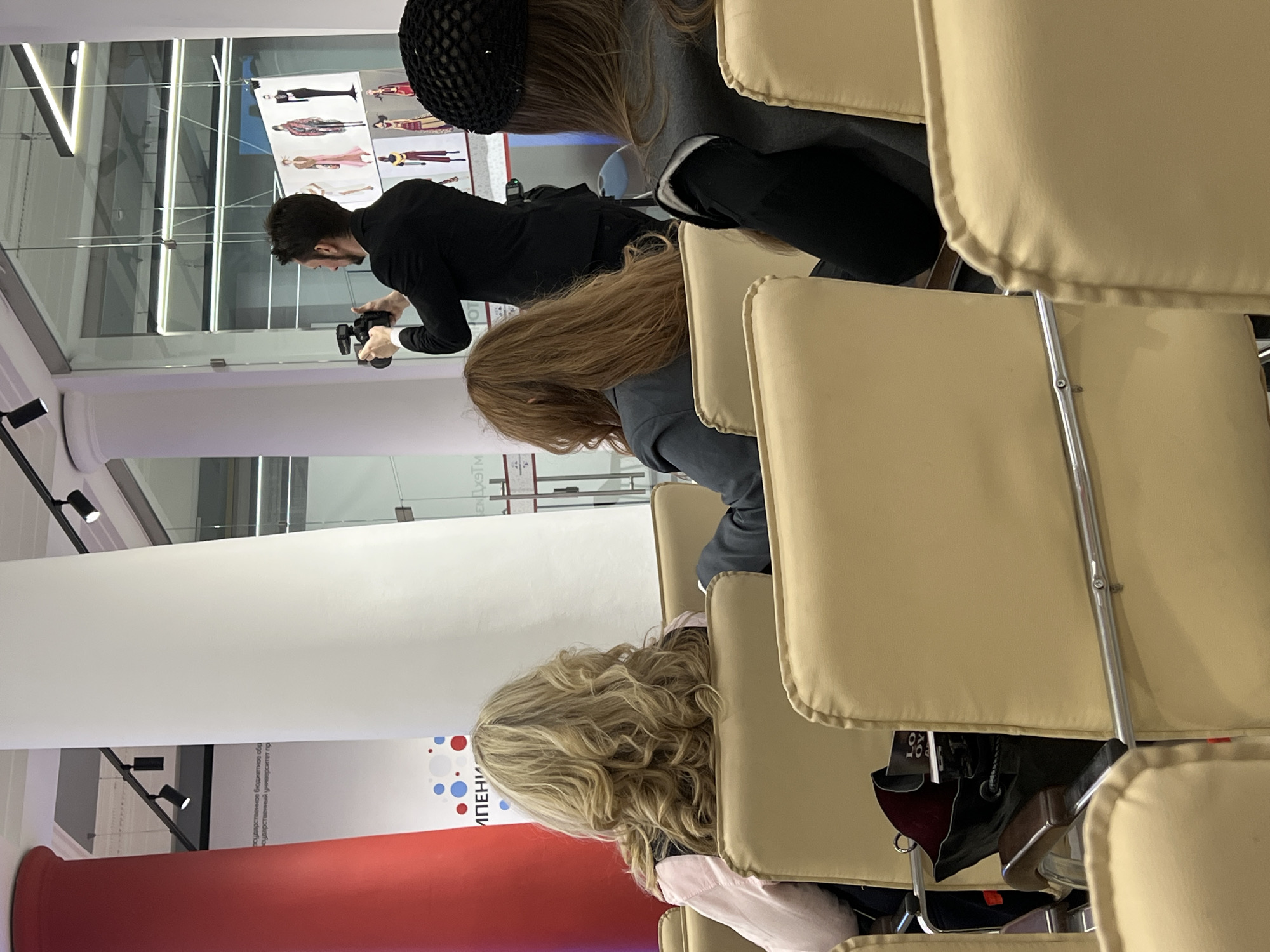 A horizontal indoor shot shows a photographer taking a photo, while the heads and backs of viewers in chairs fill the rest of the image.
The photographer, who faces left, wears a long-sleeved black shirt. He has dark hair and a beard. A professional camera is in his hands. Behind him is a wall of glass with images on the opposite side.
The viewers are seated in tan-colored chairs with silver metal frames. Three of the viewers are in the foreground and closest to the camera. One viewer in the back has her hair in a black net. The hair of the other two viewers closest to the camera are long, straight, and blonde.
A red cylindrical pillar is visible in the background with a white background behind it. An image of colorful dots is on the white wall, with lettering in Cyrillic. The city is Saint Petersburg, Russia.