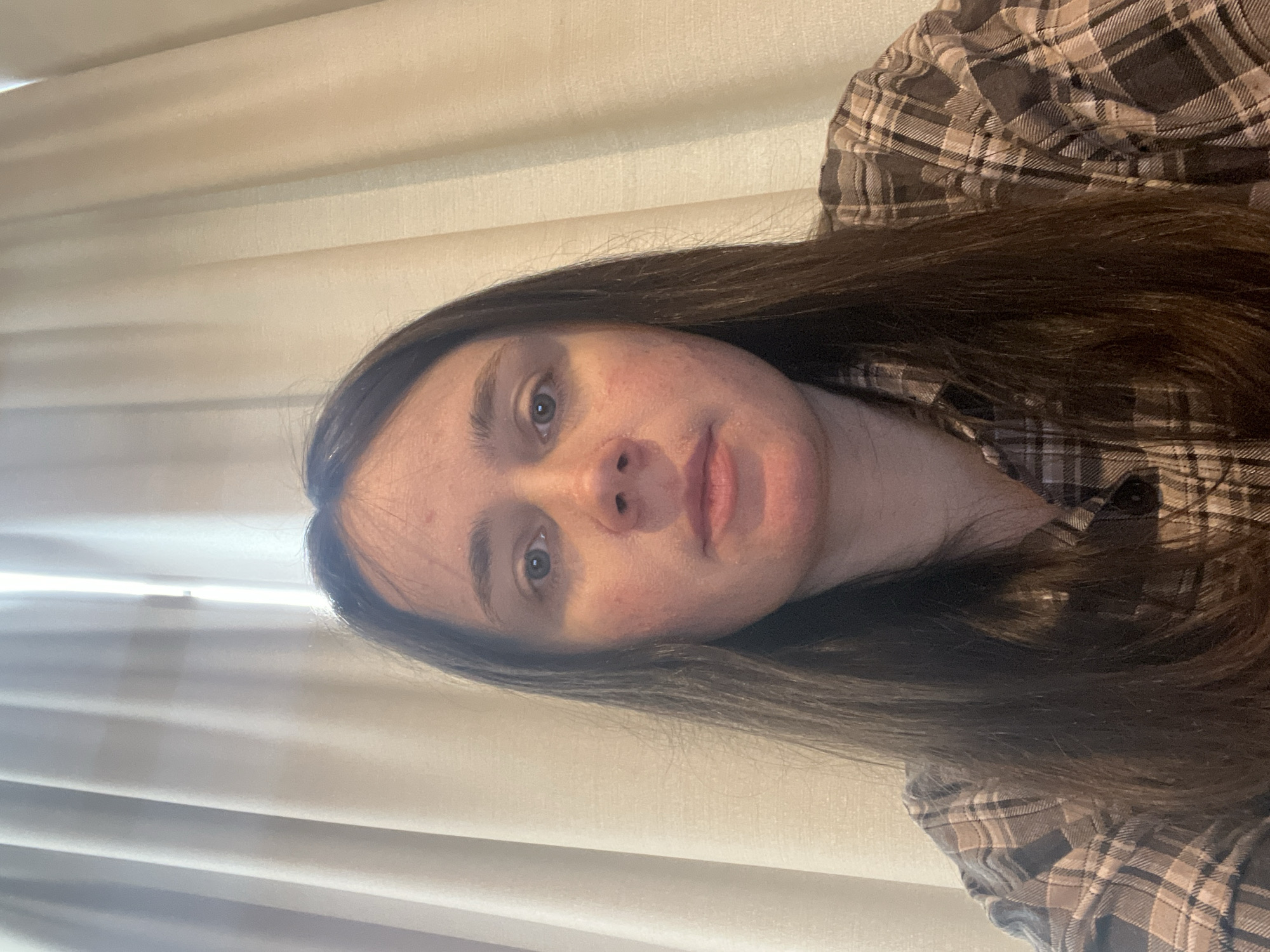 A selfie taken by a young woman with long, dark brown hair is shown. She is wearing a plaid shirt and is looking directly at the camera. The background consists of cream-colored curtains. The lighting is soft and diffused, coming from behind her. The image appears to have been taken indoors, possibly in a room. There is no discernible city or location information present in the image.