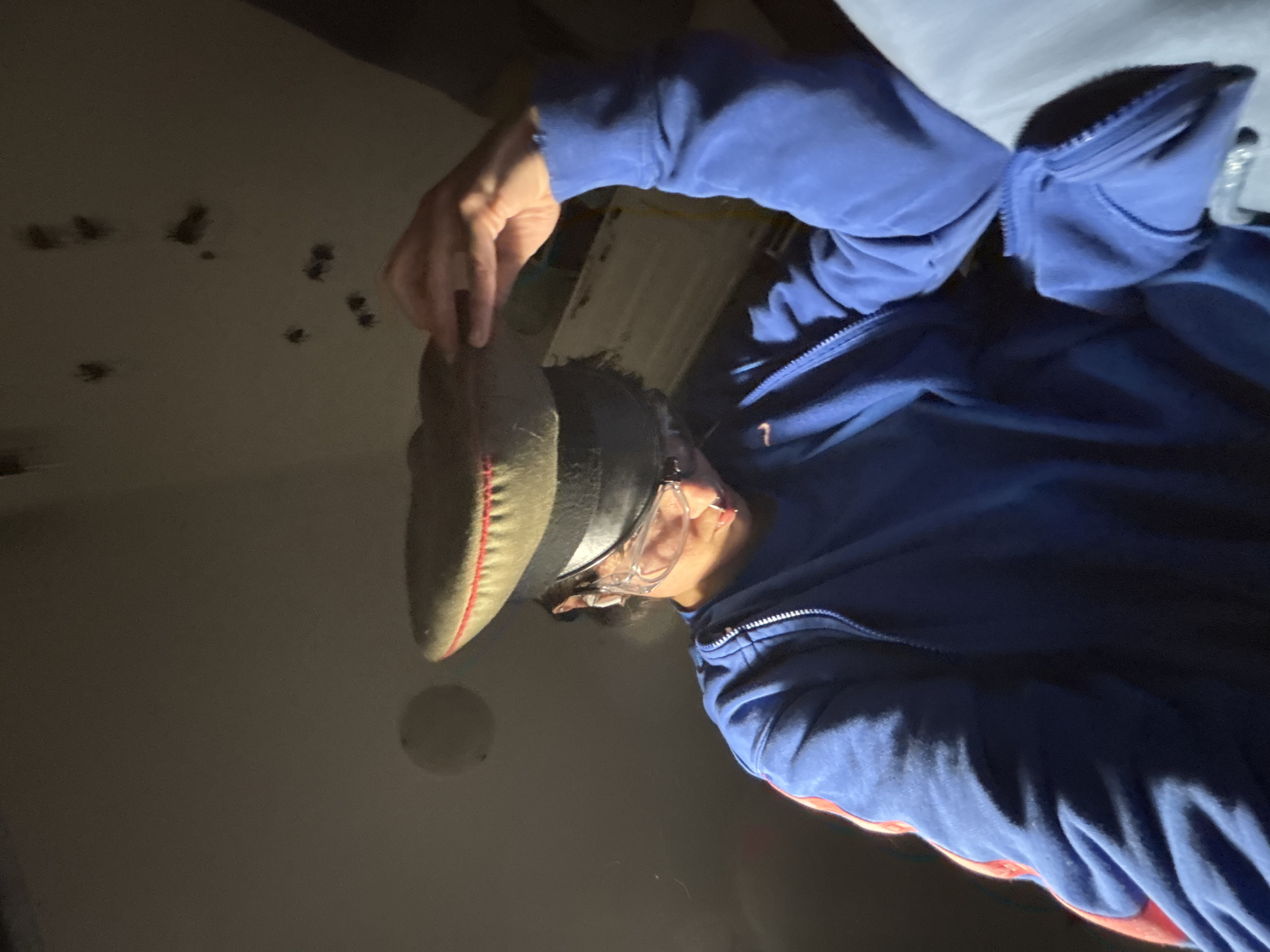 A person wearing a blue jacket, safety glasses, and holding a grey hat is standing in a room. The walls are grey. There are holes on the walls. The room appears dark. There is no clear evidence of location in Sofia, Bulgaria.