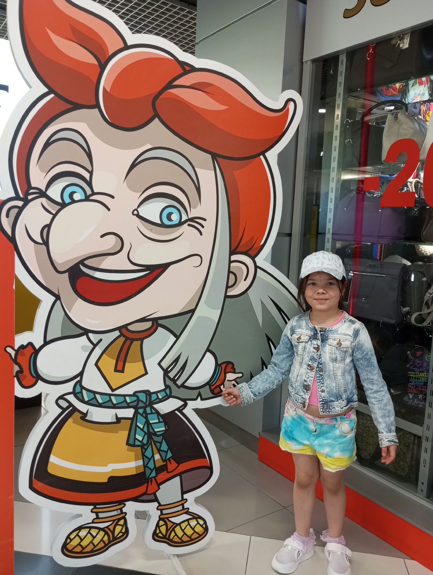 A young girl stands next to a large, cartoonish cutout of a woman with red hair, a large nose, and a traditional-looking outfit. The girl wears a light-colored baseball cap, a denim jacket, tie-dye shorts, and white sneakers with purple accents. The backdrop includes a store window filled with purses and bags, marked with a red "20%" sign. This photograph was taken in Izhevsk, Russia.