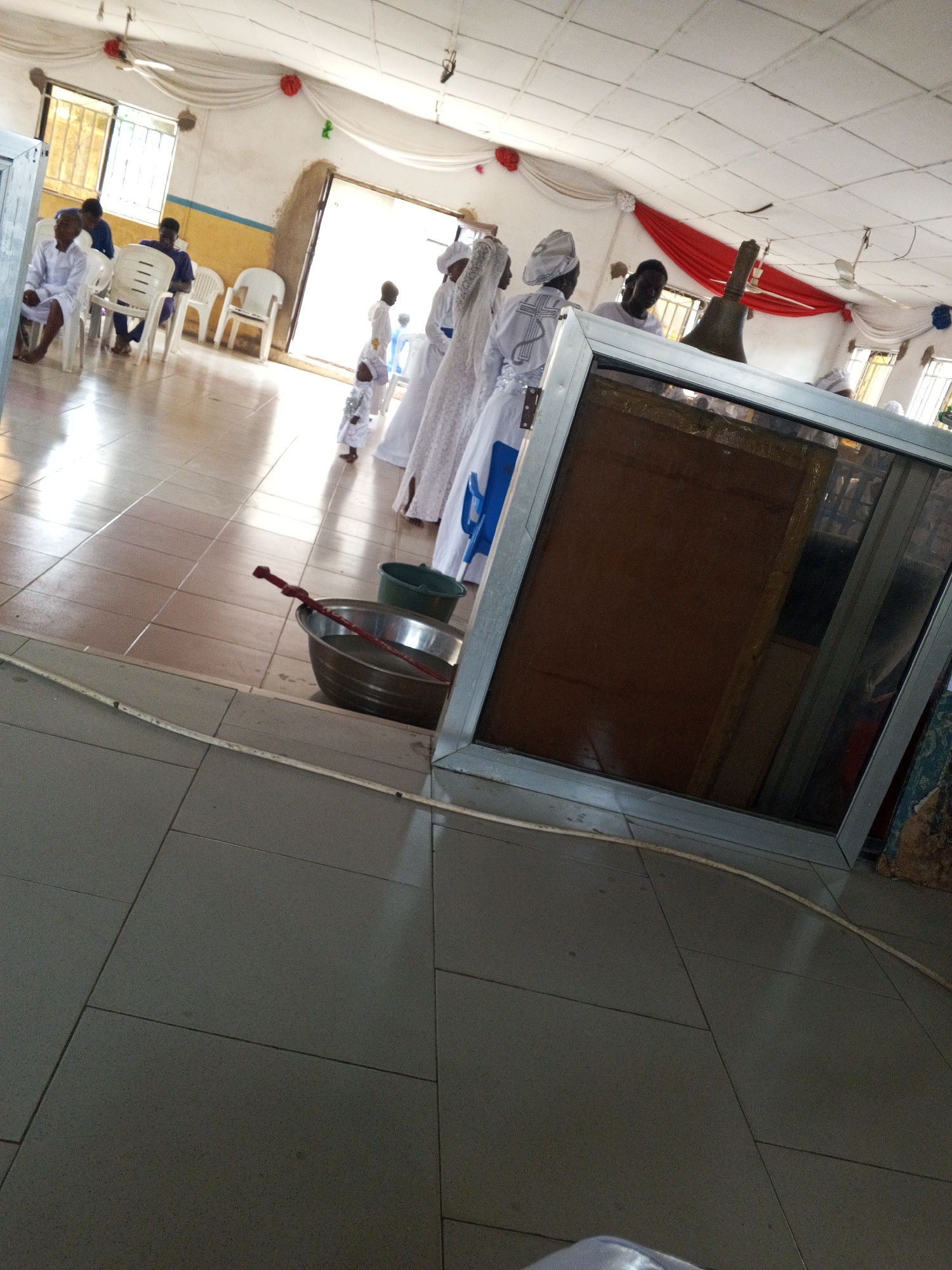 The photograph shows the interior of a building, possibly a church or religious hall. Several people are present, some dressed in white robes and head coverings. A child is also visible. There are chairs along the wall. In the foreground, there is a silver basin or container with a stick or tool inside it. A framed object is standing on the floor, obstructing part of the view. The floor is tiled. The location is not identifiable in the image.