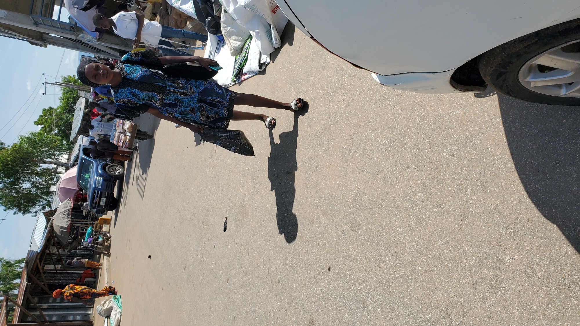 The image depicts a daytime scene in a bustling marketplace in Jalingo, Nigeria. A young woman, wearing a blue and black patterned dress and white sandals, is walking away from the camera, carrying a black bag in her right hand and holding a green phone in her left. Her shadow is cast prominently on the asphalt road. In the background, a blue pickup truck is parked, and various market stalls with awnings are visible. Several people are milling about, including a man in a white t-shirt with "11 betsson" written on it, pushing a cart laden with loaves of bread. Another woman in a colorful patterned garment stands near some stalls on the left side of the image. The environment appears to be dusty and somewhat unkempt, consistent with a busy outdoor market. A portion of a white vehicle is in the foreground on the right.