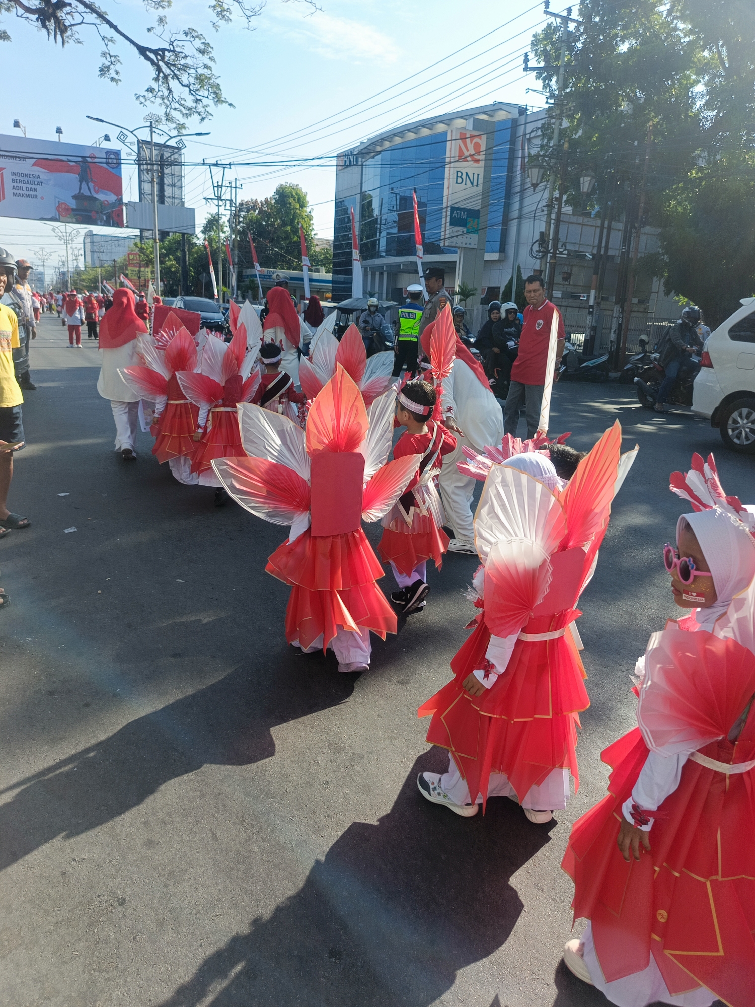 The image shows a street scene in Gorontalo, Indonesia, likely during a parade or celebration. Several children, dressed in elaborate red and white costumes resembling petals or wings, are walking in a procession. The costumes feature layered red fabric with gold trim and translucent white accents. Some of the children are wearing headscarves, and one child has pink sunglasses and face paint.
In the background, there are buildings, including a BNI (Bank Negara Indonesia) branch with a prominent blue facade and signage. A large banner with Indonesian flags and text is visible on a billboard structure. Several adults, some in casual attire and others in what appear to be uniform or ceremonial clothing, are also present. Motorcycles are parked along the side of the road, and a white car is partially visible. The sun is shining, casting distinct shadows on the asphalt road, suggesting it is daytime. The presence of police officers, one in a green vest with "POLISI" written on it, indicates an organized event possibly requiring crowd control or security. The overall atmosphere appears festive.