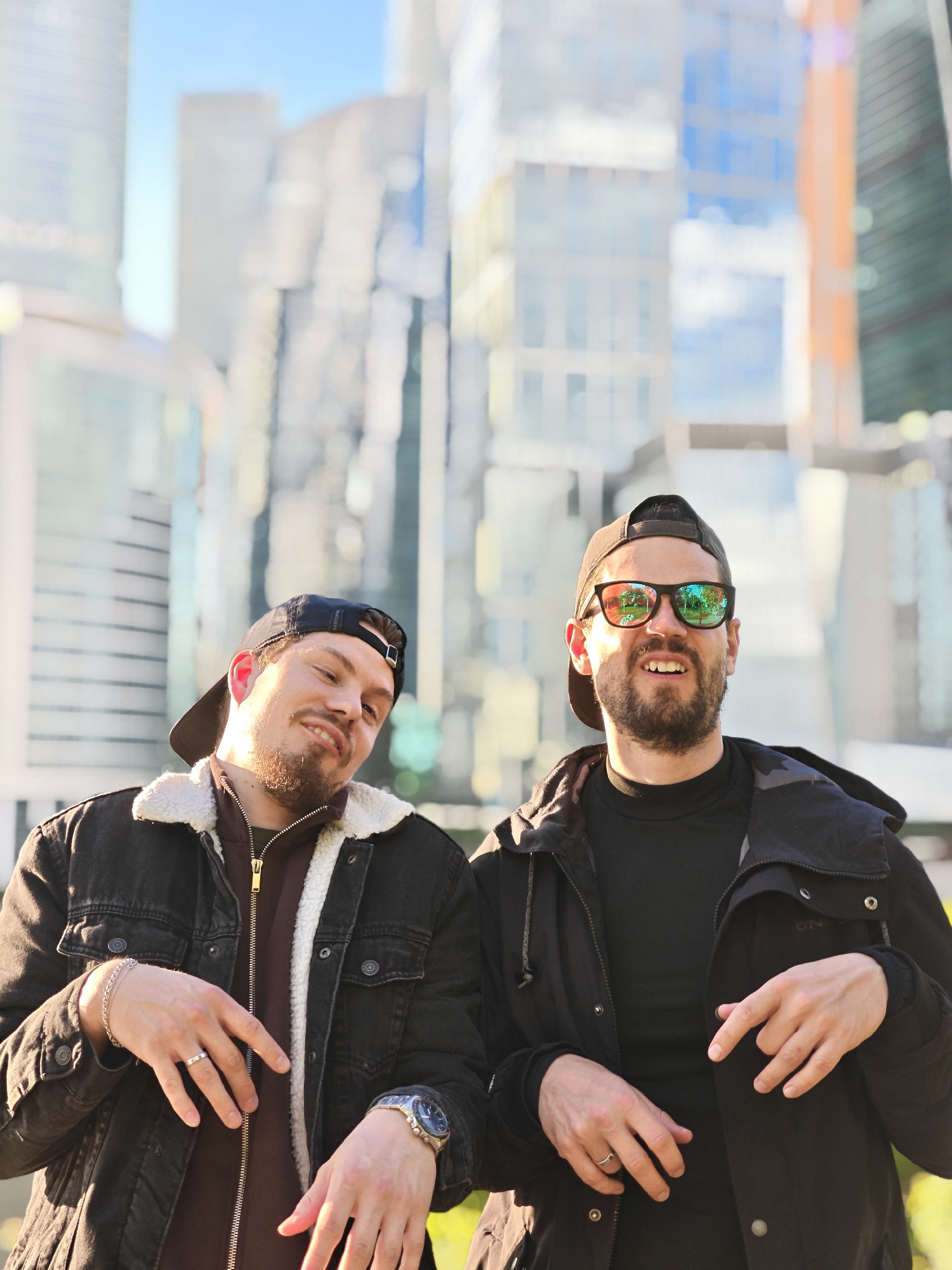 Two bearded men are posed in front of a blurred background of skyscrapers, in Moscow, Russia. The man on the left wears a black baseball cap backward, a black denim jacket with a white fleece collar, and a maroon zip-up shirt. He has a silver ring on his left ring finger and a watch on his left wrist. The man on the right wears a black baseball cap backward, sunglasses, a black jacket, and a black turtleneck shirt. He has a silver ring on his left ring finger. Both men have their hands in front of them, positioned as if holding a small object. The background consists of indistinct high-rise buildings, indicative of a modern urban environment. The overall lighting is bright, suggesting a sunny day.