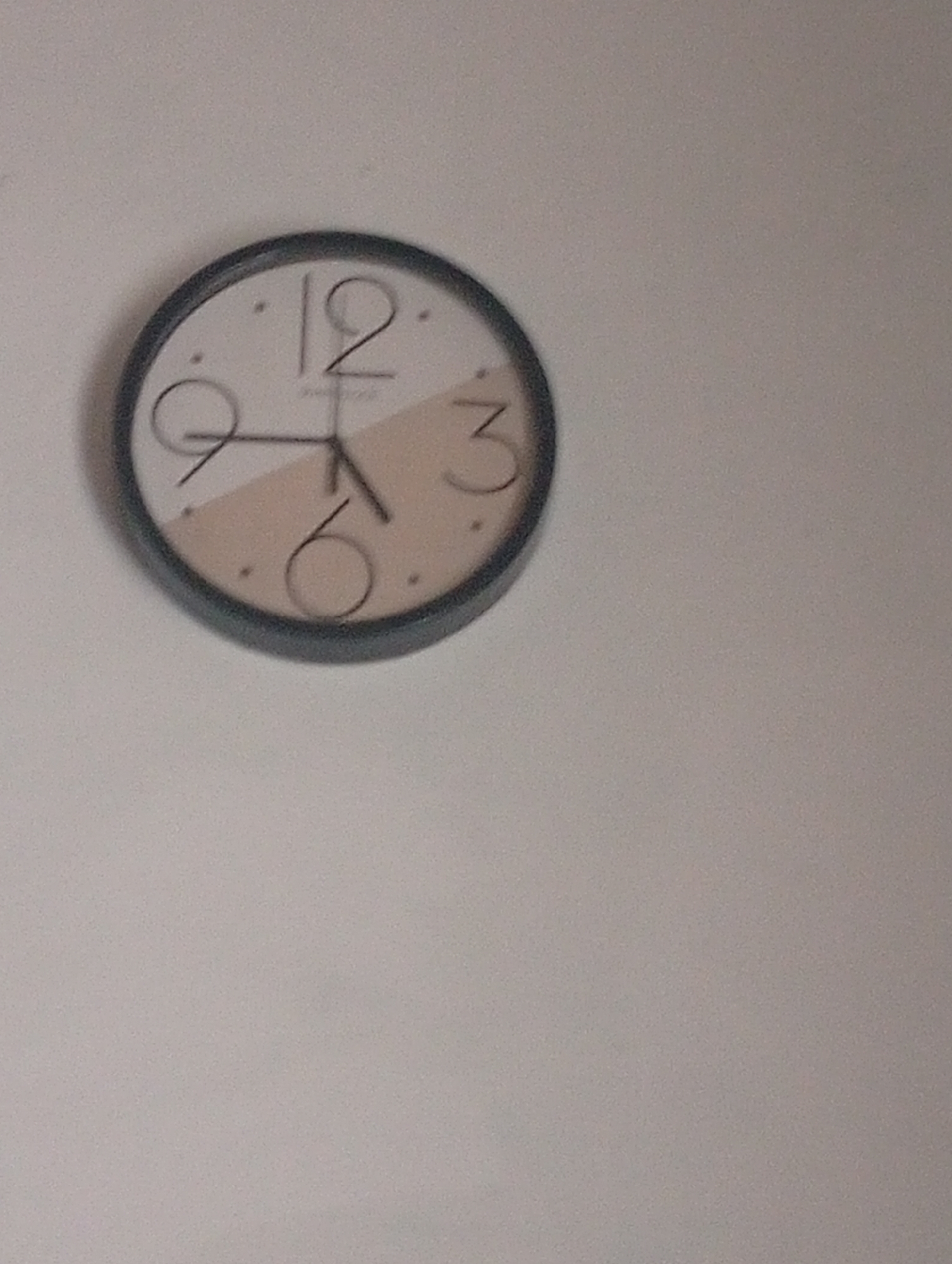 A round, analog clock hangs on a plain, light-colored wall. The clock has a dark rim and a white face divided by a diagonal cream-colored segment. The numbers 12, 3, and 6 are visible, with the 12 at the top, 3 to the right, and 6 at the bottom. The clock hands are dark and appear to be pointing close to 12:30. The 9 is also visible on the left side. The overall setting is nondescript, with no other objects or people in the frame. The image quality is somewhat grainy.