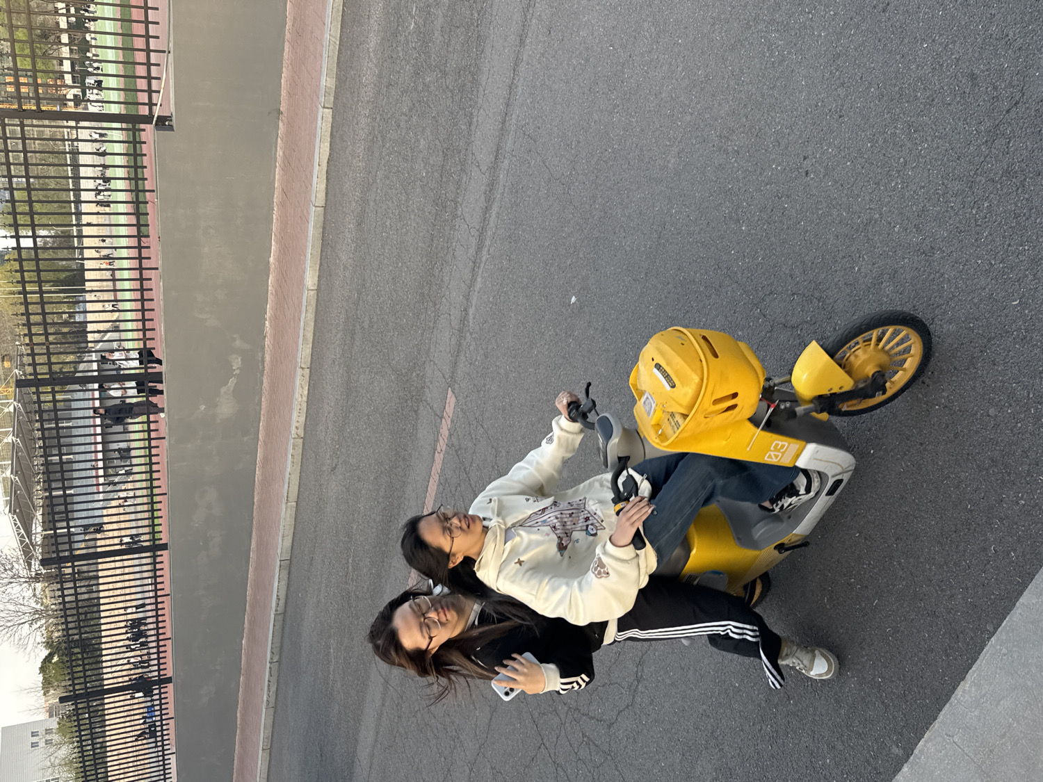 Two young women are depicted riding a bright yellow shared electric scooter on an asphalt road in what appears to be an urban or campus setting in Zhongtun, China. The image is rotated 90 degrees counter-clockwise.
The woman in the front, with dark hair and glasses, is dressed in a light-colored long-sleeved top with a graphic design, dark pants, and black and white sneakers. She holds the handlebars of the scooter, suggesting she is operating it. Seated behind her, another woman, also with dark hair and glasses, wears a black top, black athletic pants with white stripes, and light-colored sneakers. She is holding a smartphone. Both women appear to be smiling or in a pleasant mood, indicating enjoyment.
The scooter is predominantly yellow, featuring a matching yellow basket or storage unit on the handlebars. The number "83" is prominently displayed on its side, and some indistinct text or a logo is visible on the basket.
In the background, a black metal fence runs parallel to the road, behind which a sports field or track is visible with several people in the distance. A concrete wall with a brick-colored curb lies between the road and the fence. Trees and a building can be seen further in the background. The scene is illuminated by clear daylight, possibly late afternoon.