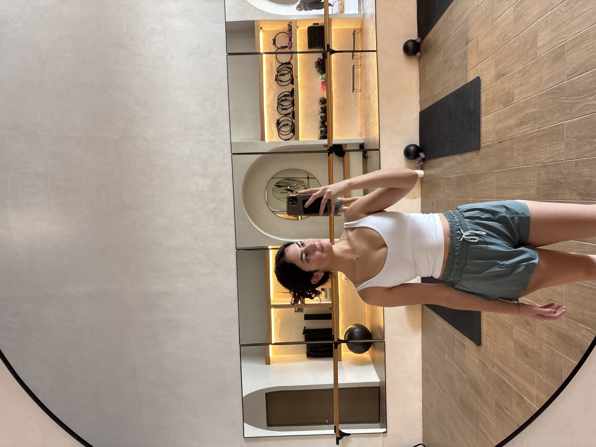 A person stands inside a pilates studio in San Bernardino Tlaxcalancingo, Mexico, and takes a mirror selfie. The person wears a white tank top and green shorts. The pilates studio has wooden flooring, black exercise mats, and a wall with large mirrors. The mirrors reflect a shelving unit containing pilates equipment, and a barre. Black pilates balls are positioned along the wall.