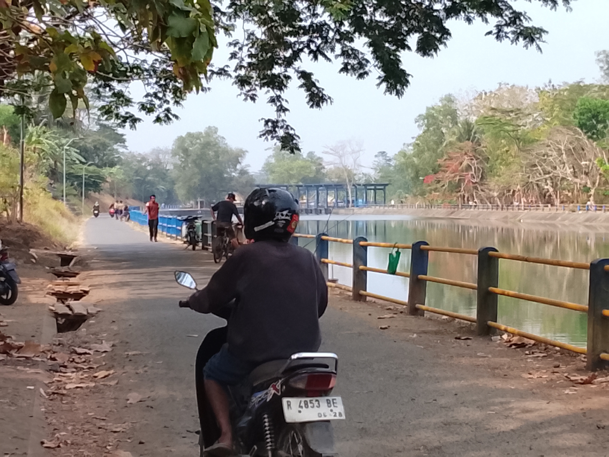 The image depicts a scene near a body of water in Kebasen, Indonesia, likely a reservoir or lake given the retaining wall and structure in the background. In the foreground, a person is riding a motorcycle from behind, wearing a dark long-sleeved shirt, blue shorts, and a helmet. The motorcycle has a license plate that reads "R 4853 BE 06-28". To the left of the motorcycle, debris and broken pavement are visible on the side of the road. Further down the road, other individuals are seen on motorcycles and walking. The environment includes trees and foliage, with a reflective body of water to the right, bordered by a yellow and blue railing. The overall atmosphere appears to be a casual outdoor setting.