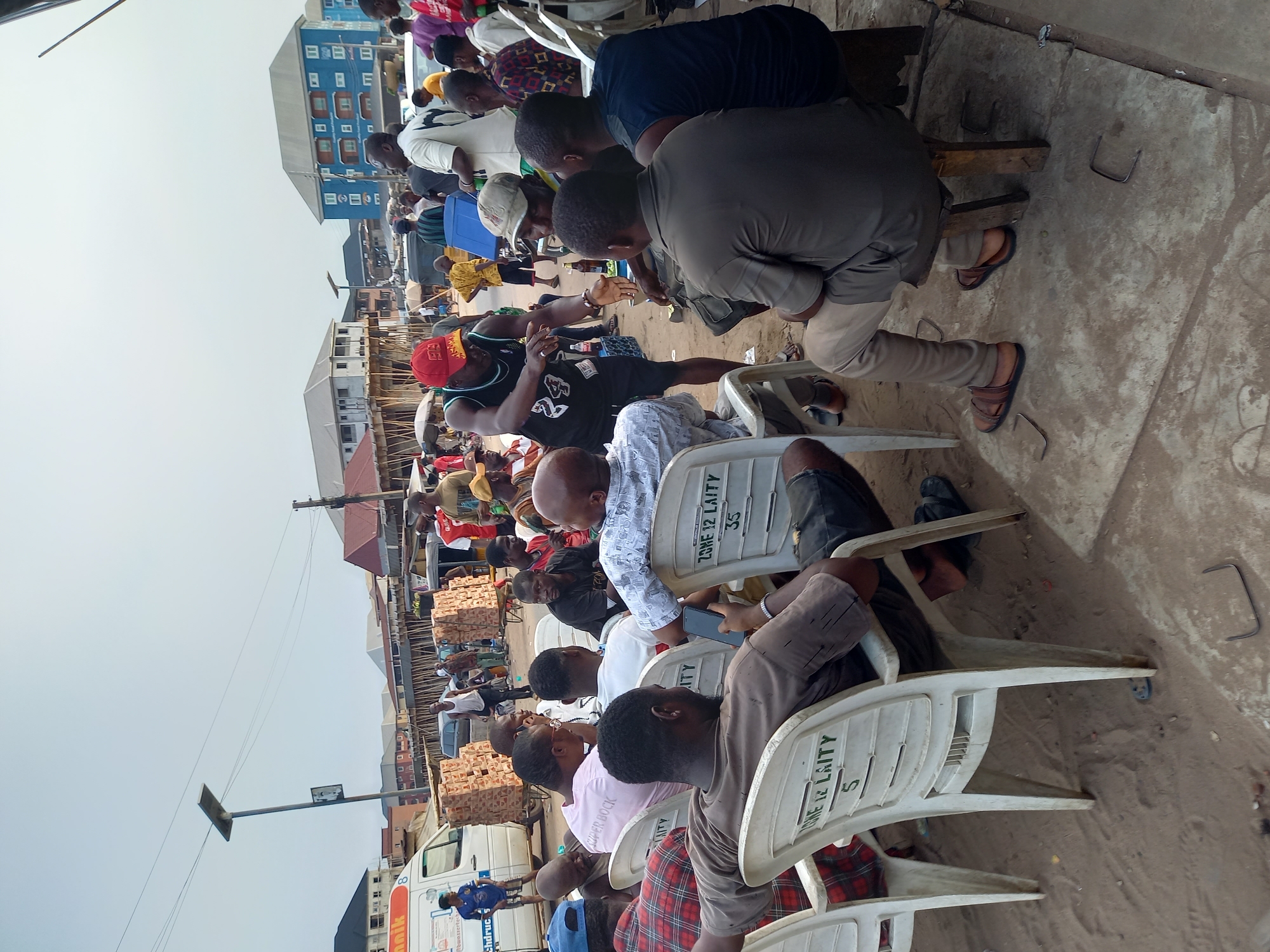A gathering of people sit in rows of plastic chairs. Many appear to be watching or listening to something outside the frame. The chairs have text on them that includes "ZONE 12 LAITY" and numbers. In the background, a building is visible and boxes are stacked behind the seated people. A white van with text is also present. The setting appears to be outdoors in a city, specifically Onitsha, Nigeria.