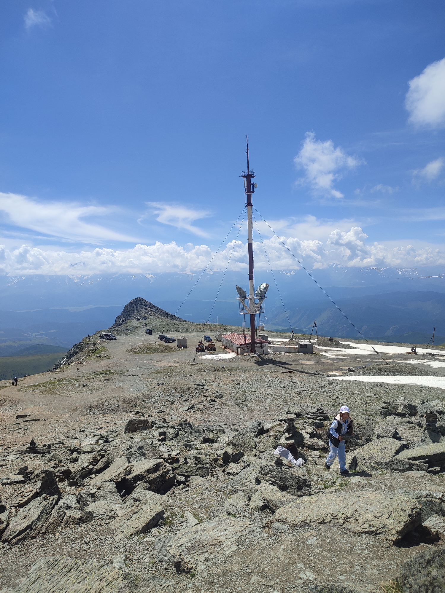 A high-angle, medium shot captures a mountainous, rocky terrain under a bright blue sky with scattered white clouds. In the mid-ground, a prominent telecommunications tower with red and white bands stands tall, equipped with satellite dishes and other equipment, connected to a small, weathered building at its base. Surrounding the tower are several parked vehicles, including what appears to be a bulldozer. Further into the distance, a jagged mountain peak rises, topped with a rocky outcrop, and beyond that, a range of snow-capped mountains stretches across the horizon under a hazy sky. In the foreground, a young person wearing a white hat, a light blue jacket, and pants walks through a rocky area, holding a piece of rock. Another person in darker clothing is visible further back on a dirt path, walking away from the camera. The ground is covered in loose rocks, gravel, and patches of sparse vegetation and lingering snow.