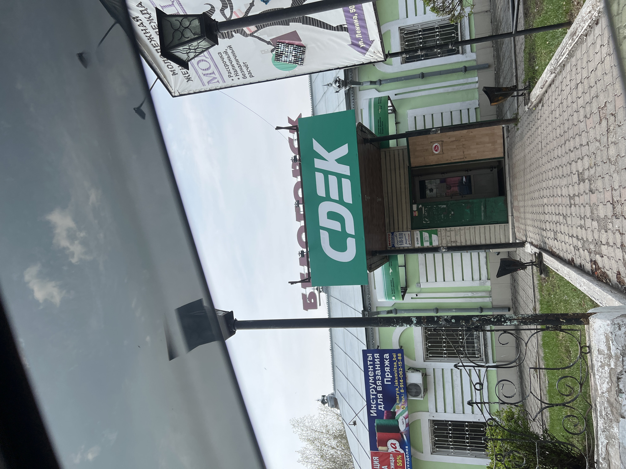 The image shows a street scene in what appears to be a small town. The main focus is a green CDEK sign, indicating the location of a CDEK office. The office building itself is light green with white trim. To the left of the CDEK sign, there are other signs including one advertising yarn and knitting supplies, and another sign that says "МОТО МОЛОДЕЖНАЯ ЖЕН", which is partially obscured. The street name on the sign is ул. Ленина, 50. A wrought iron fence runs along the sidewalk. The picture appears to be taken from inside a car, based on the partial view of the car's interior and the reflection in the window. The sky is visible in the reflection and above the buildings.