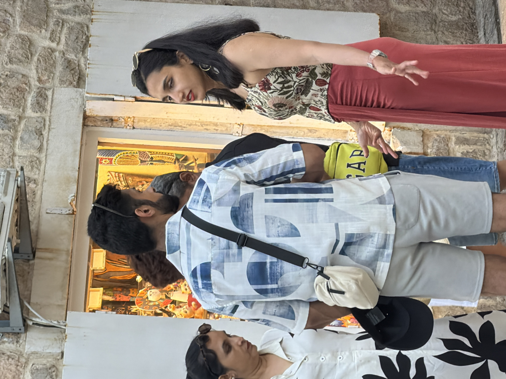 In Budva, Montenegro, three individuals are captured in a medium shot standing in front of a shop display. A man in the foreground faces away from the camera, wearing a white short-sleeved shirt with a blue abstract print, light gray shorts, and a cream-colored crossbody bag. To his right, a woman with dark hair and sunglasses on her head, adorned with a floral print top and a coral-colored skirt, engages in a conversation with him, gesturing with her hands. To the left of the man, a woman in a white dress with a black floral pattern looks down, holding a black baseball cap. The shop window behind them showcases various souvenirs, including what appear to be small figurines and textiles with intricate patterns. The background consists of a stone wall.
