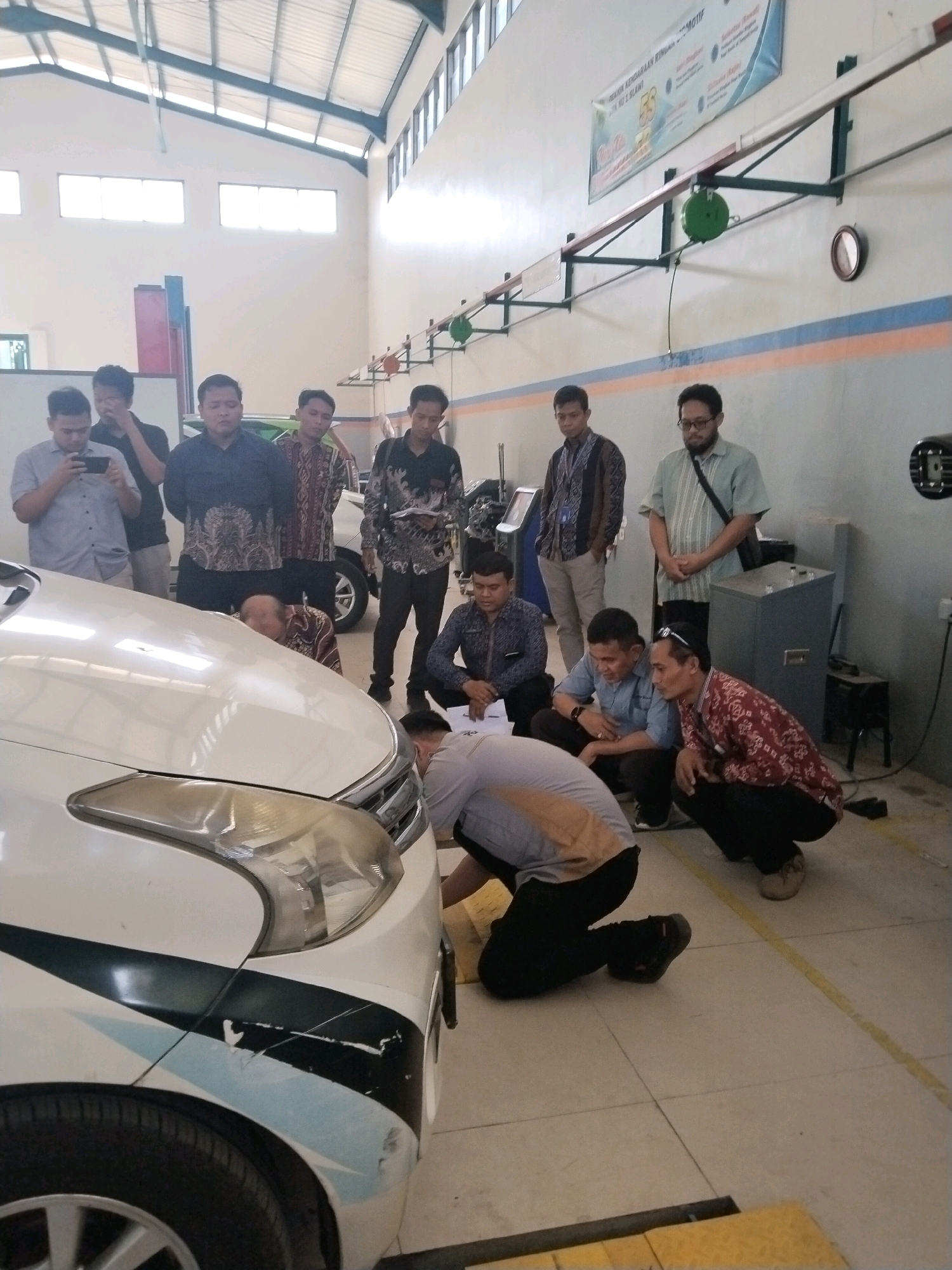 The image depicts an indoor automotive workshop, likely at a vocational school in Procot, Indonesia, given the signage. A white car is positioned prominently in the foreground, with its hood slightly ajar. Several individuals, all appearing to be adult males, are gathered around the vehicle, some standing and others in a crouched or kneeling position.
There are at least ten individuals visible. Some are dressed in casual attire, while others wear what appears to be uniforms or more formal shirts. Two individuals are focused on the engine bay of the car, one appearing to be demonstrating or explaining something. Other individuals are observing the activity, with some holding cell phones, possibly for recording or reference.
The environment is a workshop with concrete flooring marked with yellow lines. The walls are painted with horizontal stripes of blue and orange. Overhead, a fluorescent lighting system is visible, along with industrial equipment such as chain hoists mounted on a rail system. A large metal cabinet or control panel is situated against the back wall. A banner with Indonesian text and logos is affixed to the wall above the rail system, indicating it's likely an educational or institutional setting. The overall scene suggests a practical training session or demonstration related to automotive repair or inspection.