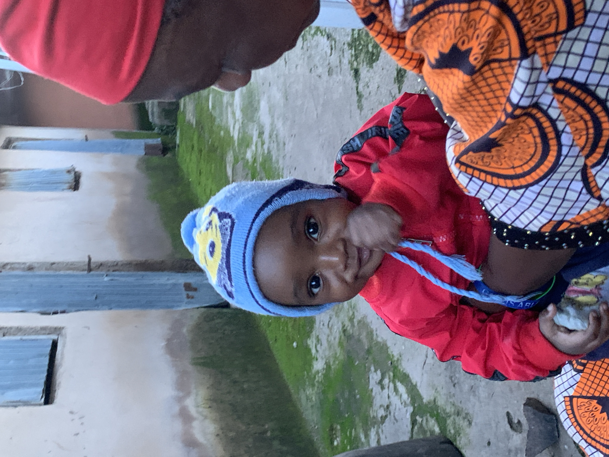 In Jos, Nigeria, a young child wearing a blue knitted hat with a yellow design and a red jacket is being held by an adult. The adult is partially visible on the right side of the frame, wearing a red headscarf and a vibrant orange and white patterned garment. The child has dark skin and is looking at the camera with a slight smile, with their hand near their mouth. A blue string from the jacket is visible. The background shows a weathered building with a blue door and a window with blue shutters, situated on a damp, mossy pathway. The environment appears to be an outdoor setting, likely a residential area.