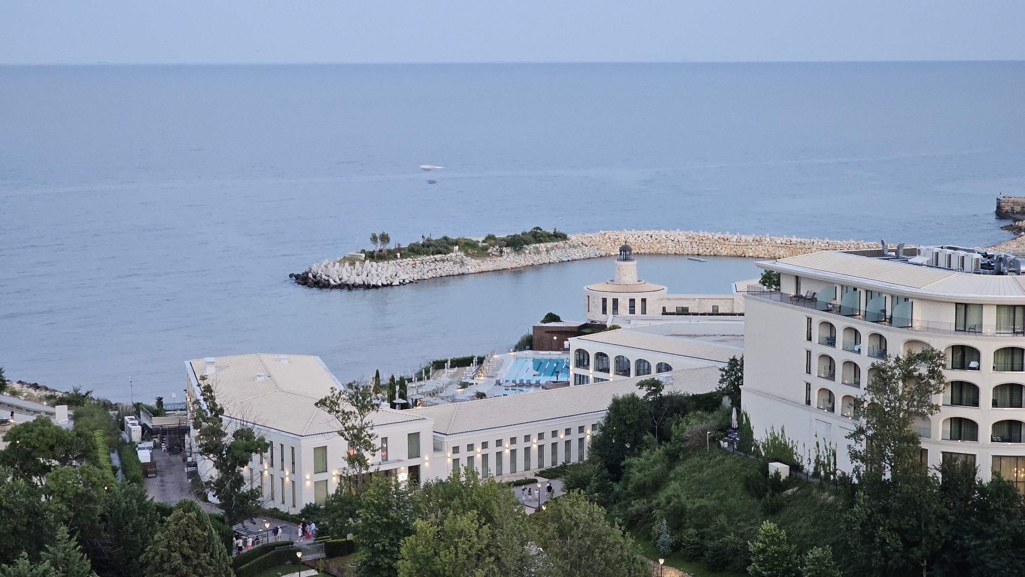 This is a high-angle, wide-shot photograph taken at dusk, showcasing a resort complex situated along the coast of Varna, Bulgaria. The scene is dominated by the expansive, calm sea in the background, with a clear horizon line where it meets the muted, overcast sky. In the middle ground, a U-shaped breakwater, constructed from large, angular concrete tetrapods and a segment of rough-hewn stones, encloses a calm bay. Within the bay, a small, rocky island with sparse vegetation and a few trees is visible, along with a lighthouse-like structure.
The resort itself is comprised of several white, modern buildings with cream-colored roofs. The most prominent building on the right is a multi-story, curved structure with numerous arched balconies. Another long, rectangular building with a colonnaded facade stretches across the middle ground. A bright blue swimming pool with lounge chairs is situated between these structures. Lush green trees and landscaping surround the buildings, creating a contrast with the architectural elements. A paved walkway or road is visible on the left side of the frame, with a few people walking and some vehicles present. The lighting suggests it is early evening, with artificial lights illuminating some of the buildings and pathways.