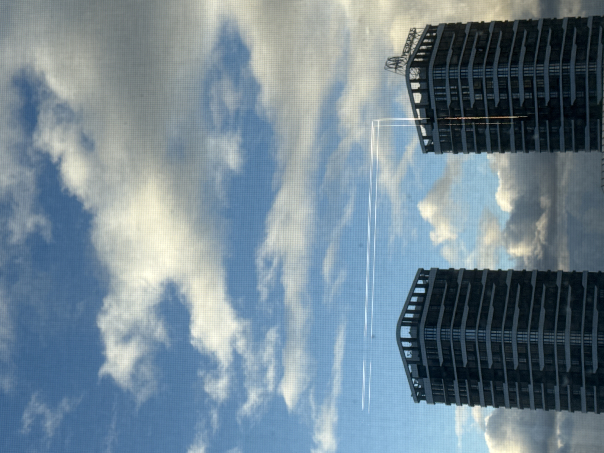 The image shows a view of the sky with clouds and a contrail, reflected in a window. Outside the window, partially visible on the right side, are two buildings. The top of one building displays the name "MERIDIAN" in large letters. Based on this detail, the image likely depicts a reflection of the "Meridians" residential complex located in Krasnoyarsk, Russia. The buildings appear to be modern high-rises with a repeating pattern of balconies or architectural protrusions. The sky behind the buildings is overcast, with visible contrails. The window through which the view is captured has a screen mesh.