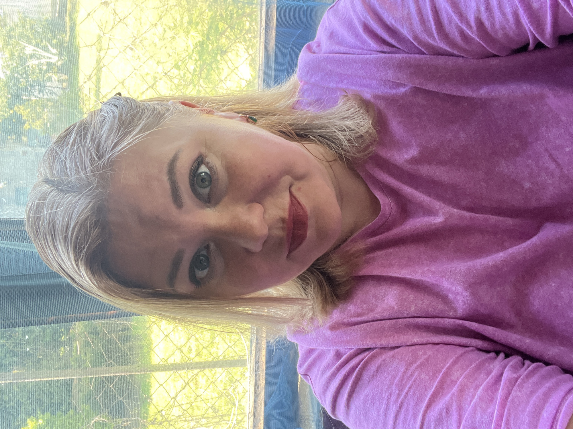 A blonde-haired woman with fair skin and green eyes is posing for a selfie. She has red lipstick on. She is wearing a pink t-shirt. Behind her is a window with a screen. Through the window, vegetation is visible. The location is unknown; there is no specific information in the image that definitively identifies the city of Vesyoloye, Russia.