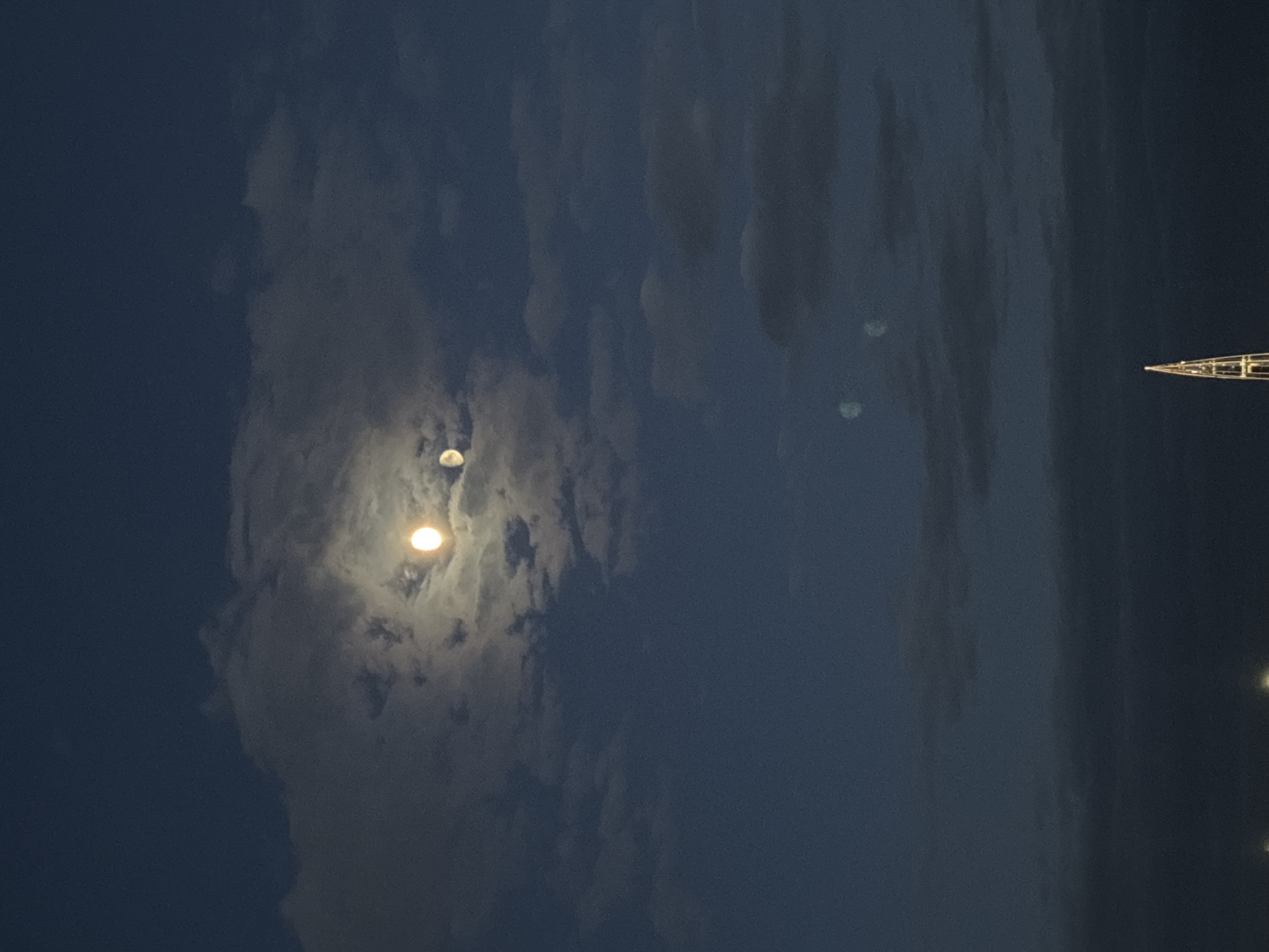 The image shows the night sky with scattered clouds and the moon visible in Saint Petersburg, Russia. The moon is partially obscured by the clouds, with a brighter glow appearing below a dimmer, secondary reflection. A radio tower is visible in the upper right of the image and what appears to be a street light is present at the bottom of the image. The sky is a dark blue color, and the clouds vary in shades of gray and white.