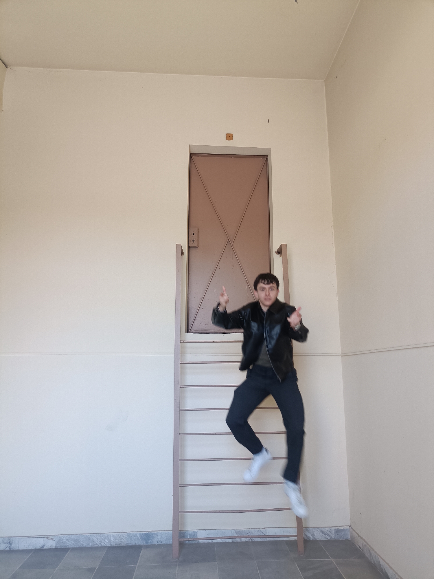The image captures a young man in mid-air, seemingly jumping or floating, in an indoor setting. He is dressed in a black leather jacket, a dark t-shirt, dark trousers, and white sneakers, which appear blurred due to motion. His hands are raised with thumbs pointing upwards, and he has a playful, energetic expression while looking towards the viewer.
The background features a tall, plain cream-colored wall. A prominent element is a closed brown metal door, positioned unusually high on the wall. This door is accessed by a matching brown metal ladder with horizontal rungs, which extends from the floor up to the doorframe. The floor is tiled with alternating light and dark gray rectangular tiles, and a light-colored marble or stone skirting board runs along the base of the wall. The room appears brightly lit, suggesting daytime conditions indoors. The arrangement of the elevated door and ladder gives the location a distinctive and unconventional appearance, possibly an institutional building or a uniquely designed interior. No text is visible in the image.