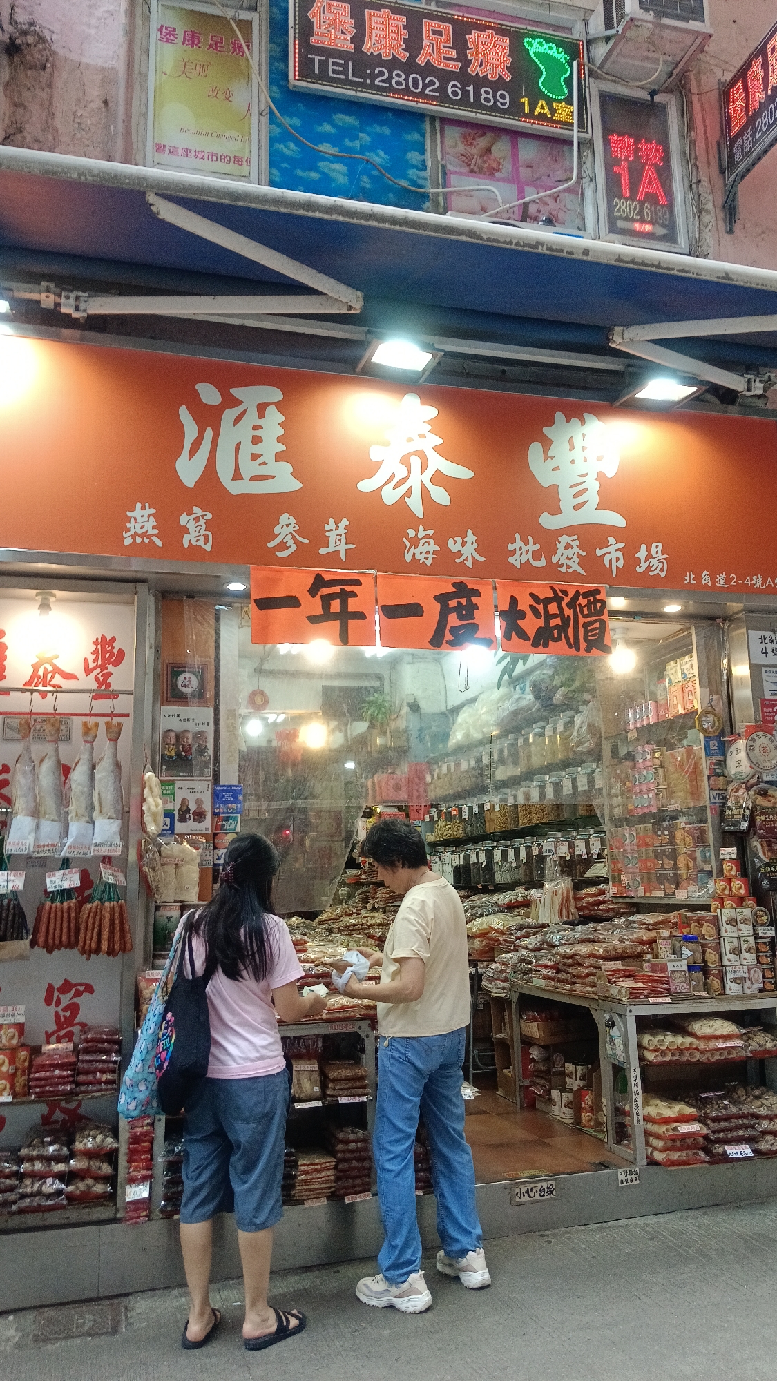 In Hong Kong, China, two women are standing in front of a shop that sells dried seafood, herbs, and medicinal ingredients. The shop's facade features a large orange banner with white Chinese characters, and above that, smaller orange banners with black Chinese characters. One of these banners reads "一年一度大減價," which translates to "Annual Big Discount." The shop interior is visible through a glass window, filled with shelves stacked with various packaged food items and jars. Hanging outside the shop on the left are strings of dried sausages and what appear to be dried fish. One woman, wearing a pink t-shirt and blue capri pants, is holding a blue patterned bag on her shoulder and looking towards the shop. The second woman, wearing a beige t-shirt and blue jeans, is facing the shop and holding a small white object in her hands, possibly a product she is examining or purchasing. Both women are wearing casual footwear. Above the shop, illuminated signs are visible, including a red neon sign with Chinese characters and a phone number. The street setting suggests an outdoor marketplace or commercial street.