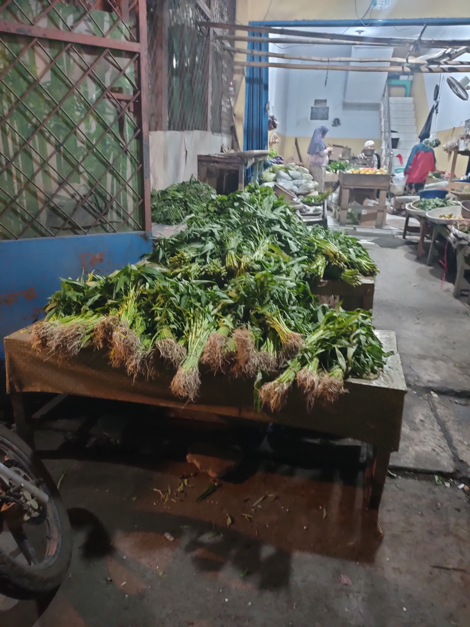 The image depicts a nighttime scene at a street-side market, likely in Subang, Indonesia. A large display of harvested water spinach (kangkung) is prominently featured on a wooden stall. The vegetables are piled high, with their roots exposed, suggesting they have just been brought to market. In the background, several individuals are visible, some wearing traditional Islamic headwear. One person appears to be attending to the produce, while others are engaged in activities related to the market. The overall environment suggests a bustling, open-air marketplace. A motorcycle is partially visible in the foreground, indicating its presence at the scene. The ground appears to be wet, possibly from recent rain or cleaning.