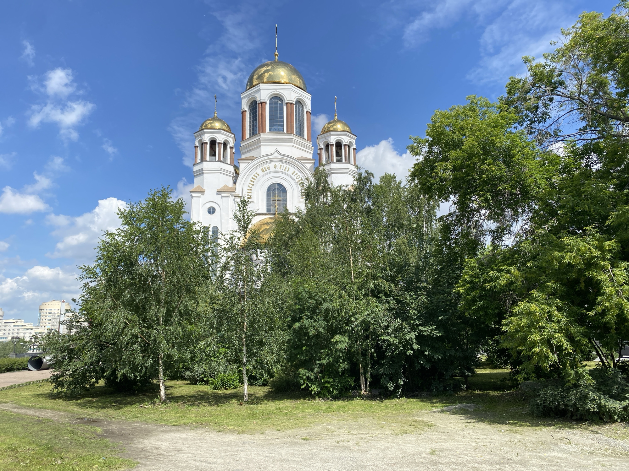The image shows the Church on the Blood in Yekaterinburg, Russia, on a sunny day with a blue sky and scattered clouds. The church is a prominent white building with golden domes and crosses. It is surrounded by lush green trees and foliage, with a gravel path and grassy area in the foreground. In the background, a modern urban landscape with tall buildings is visible. The scene depicts a peaceful and serene environment, suggesting a tourist or recreational setting.