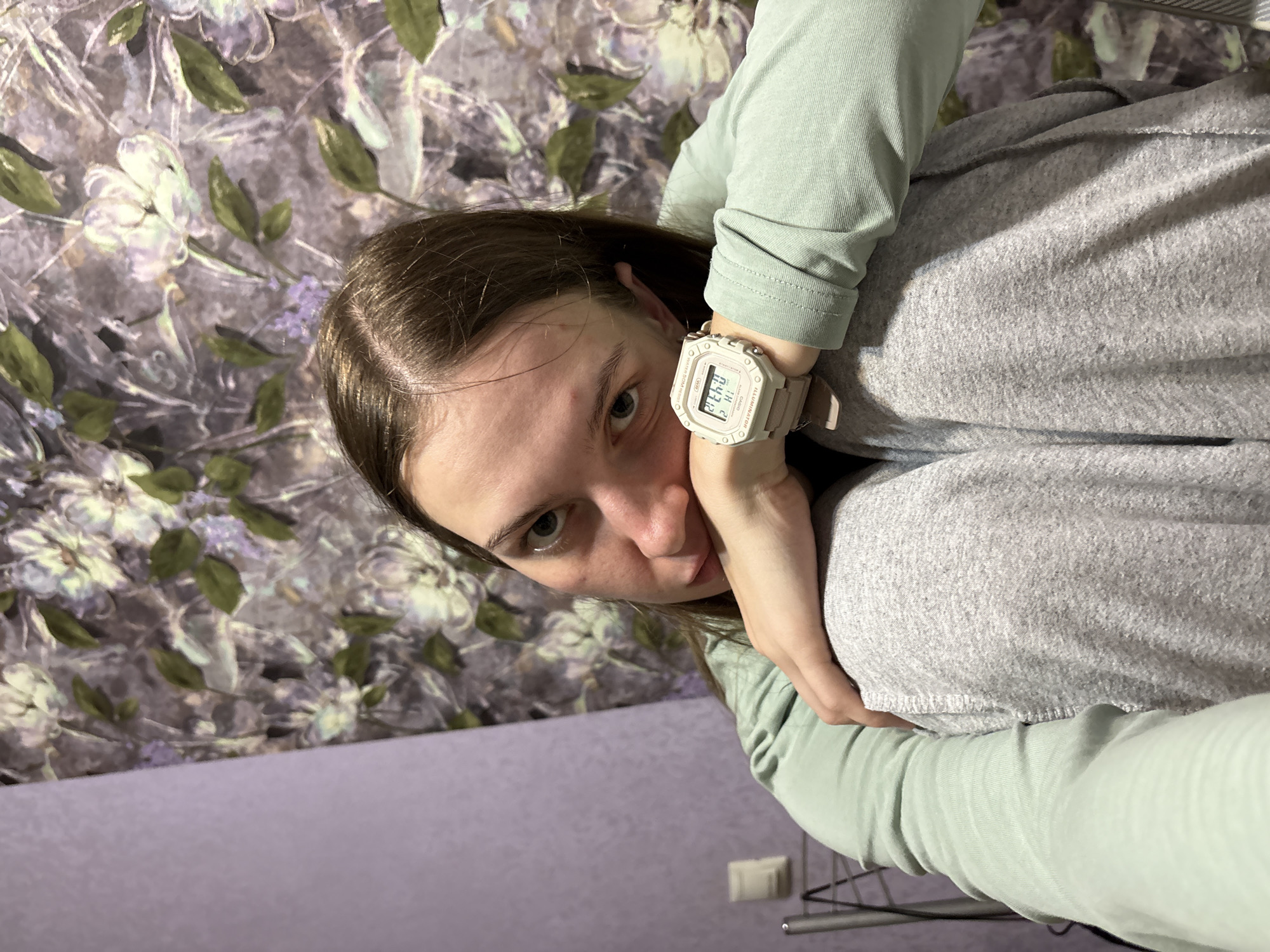 A young Caucasian female, appearing to be in her late teens or early twenties, is seated with her knees drawn up and her head resting on her arms. She has long brown hair and is looking directly at the camera. She is wearing a light green long-sleeved shirt and gray sweatpants. On her left wrist, she wears a white digital watch. The background consists of a floral wallpaper in shades of purple, white, and green, and a plain lavender wall to the left. The image appears to have been taken indoors. The timestamp on the watch shows "0:47". This scene is not identifiable as being in the city of Perm, Russia.