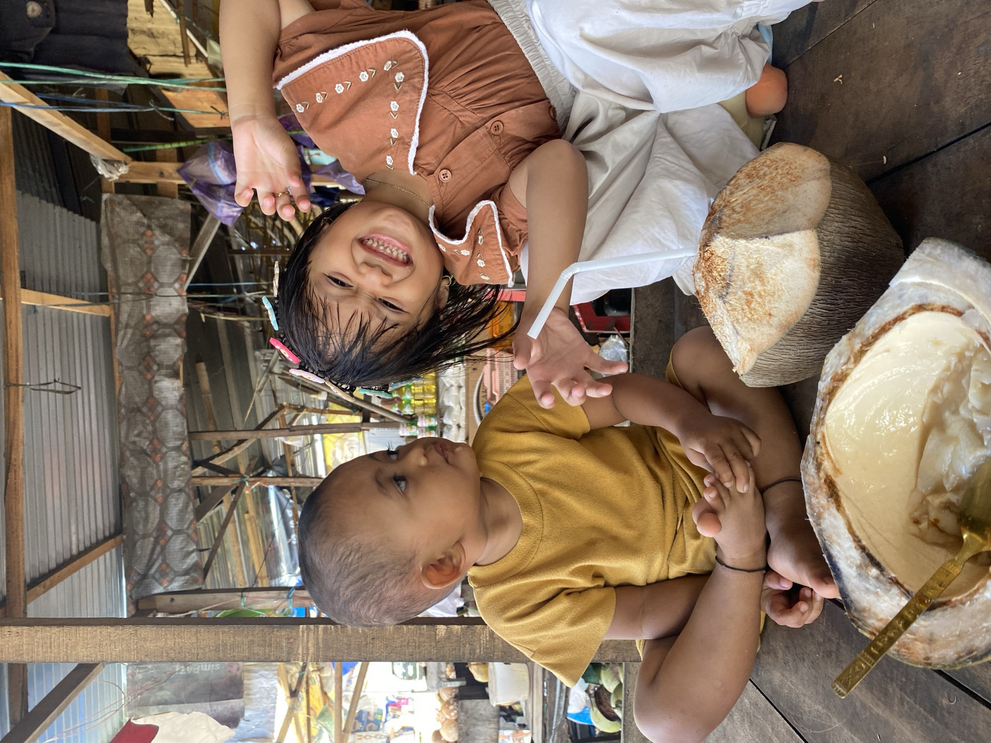 Two children are seated on a wooden surface. The child on the left is a toddler wearing a yellow t-shirt, facing right and looking towards the other child. The child on the right is a young girl wearing a brown shirt and light-colored pants, leaning back with her mouth open in a smile and her arm extended. Both children are near a halved coconut with a spoon inside. The setting appears to be an outdoor market stall, with wooden structures, a corrugated metal roof, and various goods visible in the background, suggesting a location in Ta', Indonesia.