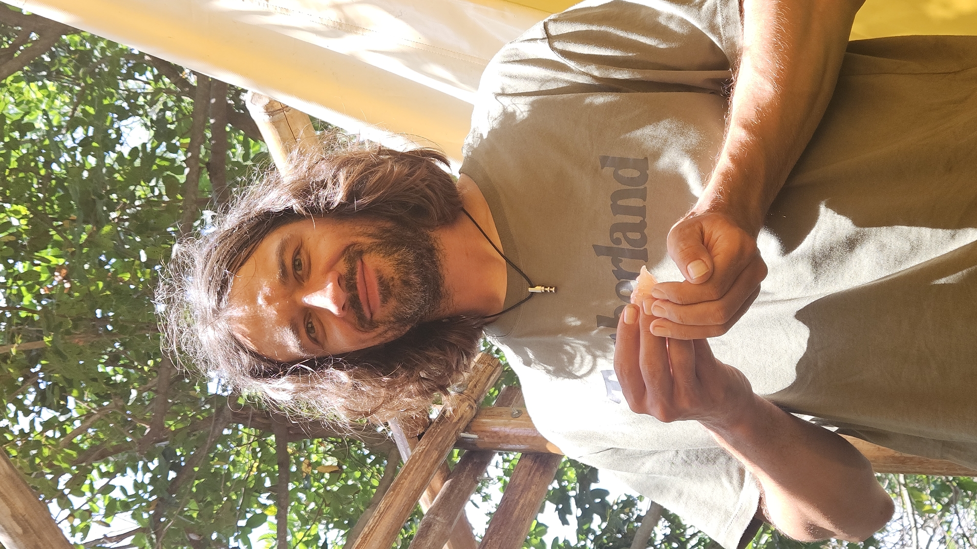 A man with long brown hair and a beard is standing outside in Nha Trang, Vietnam. He is wearing an olive-green t-shirt with the word "Timberland" written vertically down the front. He has a necklace with a silver pendant. He is holding something small and white in his hands. There are trees and leaves in the background, as well as some bamboo scaffolding. The lighting suggests it is daytime, with sunlight dappling through the trees.