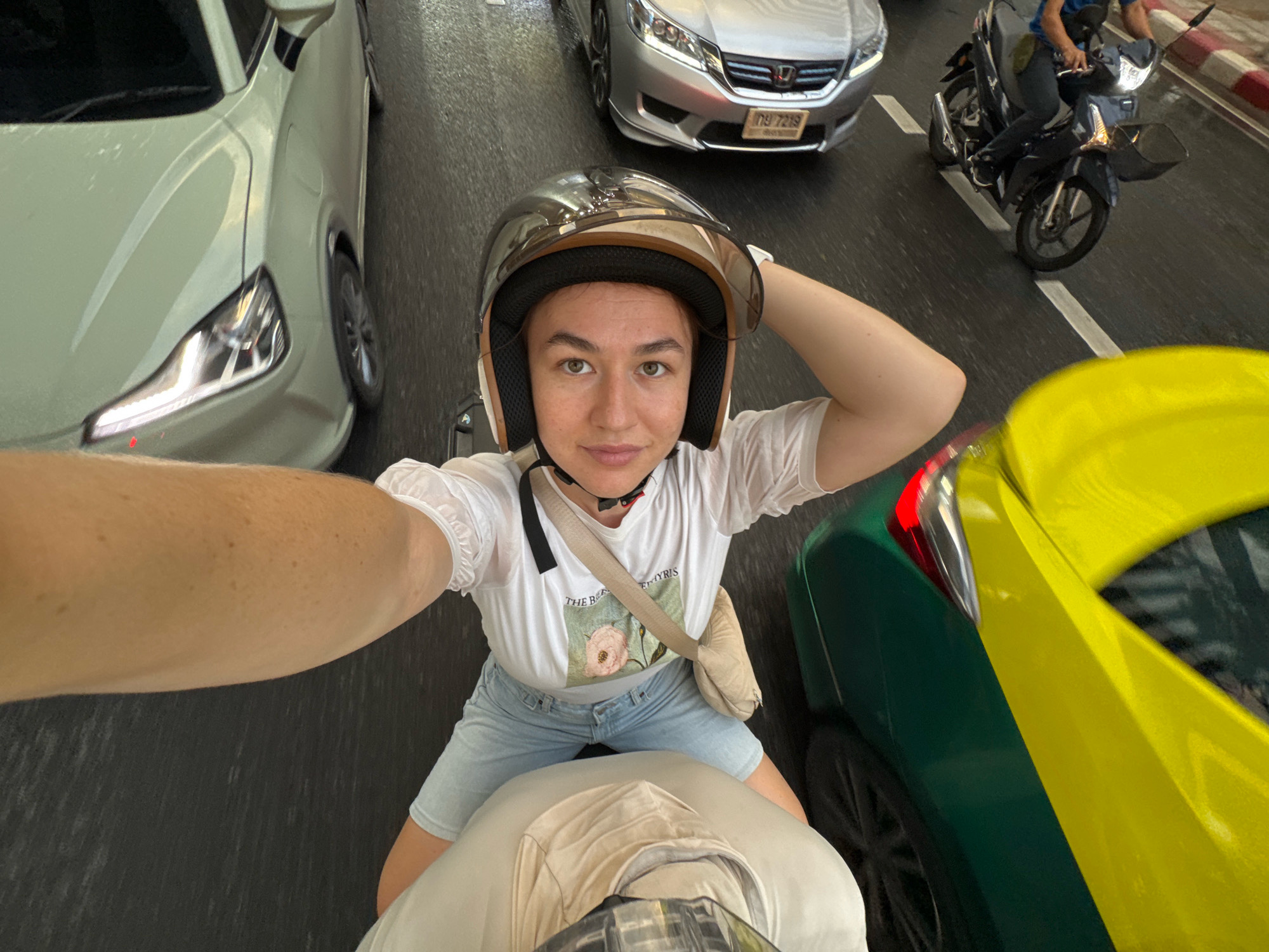 In Bangkok, Thailand, a person wearing a helmet is on the back of a motorcycle, taking a selfie. The person is dressed in a white t-shirt and light blue denim shorts. A beige crossbody bag is slung over their shoulder. The motorcycle is in traffic, with other vehicles visible, including a white car to the left, a silver sedan in the background, and a yellow and green vehicle to the right. The road is wet, suggesting recent rain. The image captures the bustling street scene typical of Bangkok.