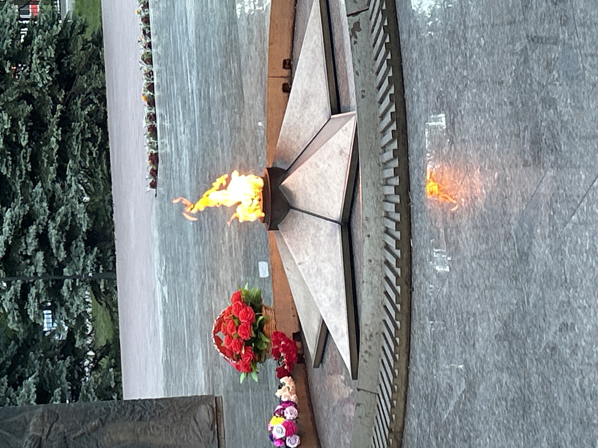 The image shows a monument in Tolyatti, Russia, featuring an eternal flame. The flame burns atop a grey, five-pointed star structure. Flowers are arranged at the base of the star. A concrete surface surrounds the monument, with trees visible in the background. A reflection of the flame can be seen on the wet concrete.