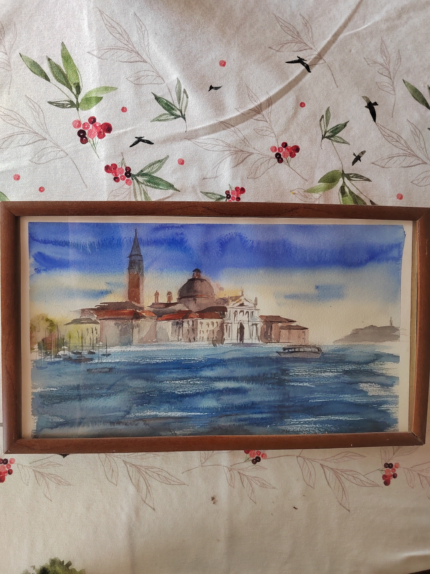 The image shows a framed watercolor painting of a cityscape with water. The painting depicts buildings with a tall bell tower and a dome, situated along a body of water. Boats are visible on the water. The sky is painted in shades of blue and yellow. The painting is framed in a wooden frame. The painting is placed on a white tablecloth with a pattern of green leaves and red berries, and silhouetted birds. The question mentions the city of Бярэзінскі сельскі Савет, Belarus; however, the painting itself does not depict this location.