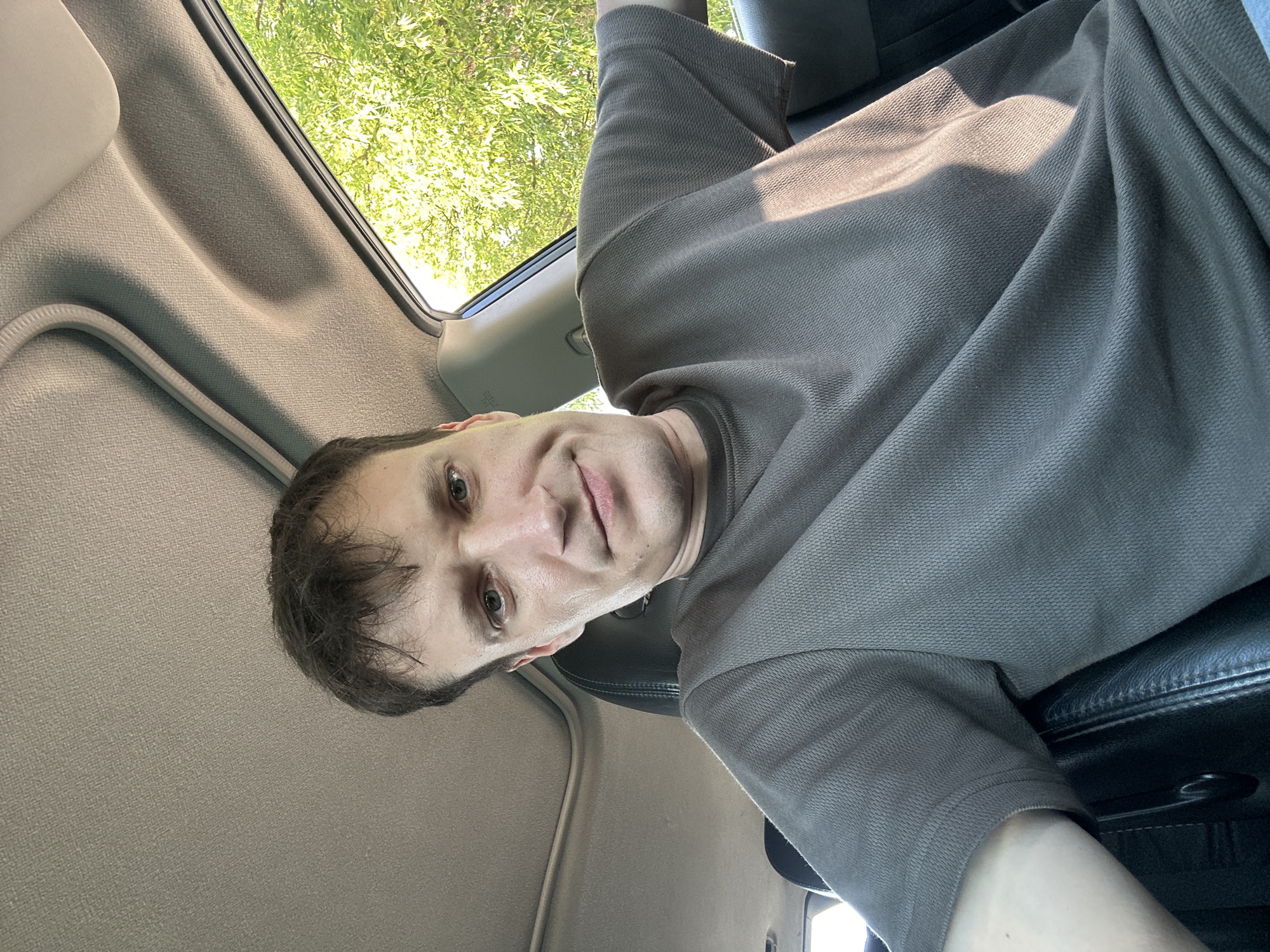 A man is sitting in the passenger seat of a car, looking at the camera. He has short, dark hair and light green eyes. He is wearing a grey t-shirt. The car's interior is visible, including the headliner and a sunroof. Sunlight streams in from the right side, illuminating the man and the car's interior. Outside the car, green trees are visible. The location is not specified as Makiv, Ukraine, nor is it implied by the image.