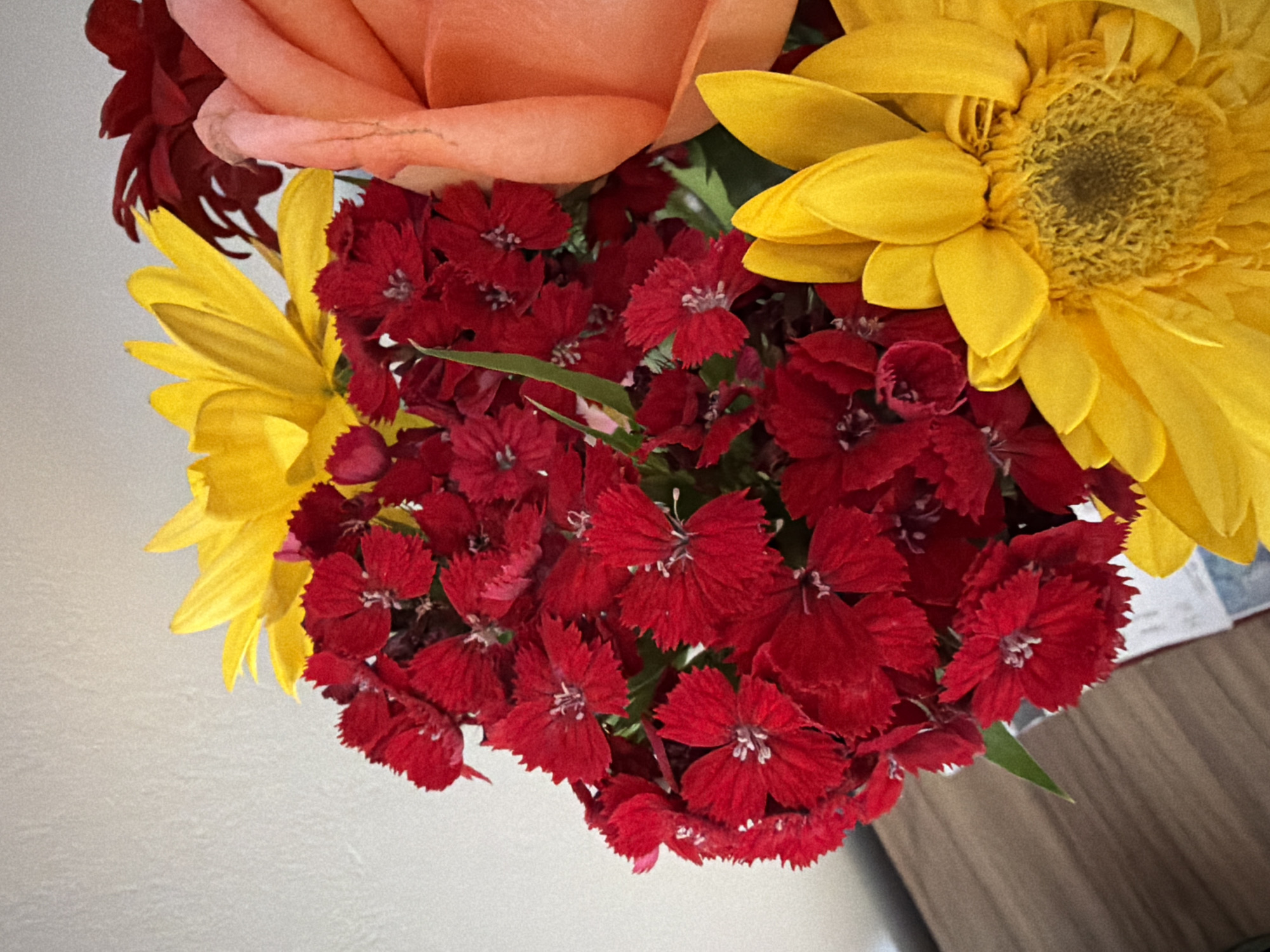 The image shows a close-up of a bouquet of flowers. The bouquet contains a peach-colored rose, two yellow sunflowers, and a cluster of small, red flowers with fringed petals. The flowers are arranged together, creating a dense, colorful display. The background is partially visible, showing a white wall on one side and a brown wooden surface on the other. The image is taken from an overhead perspective, looking down at the arrangement.
The location in the city of Catania, Italy is not provided in this image and cannot be determined from the context of the image.