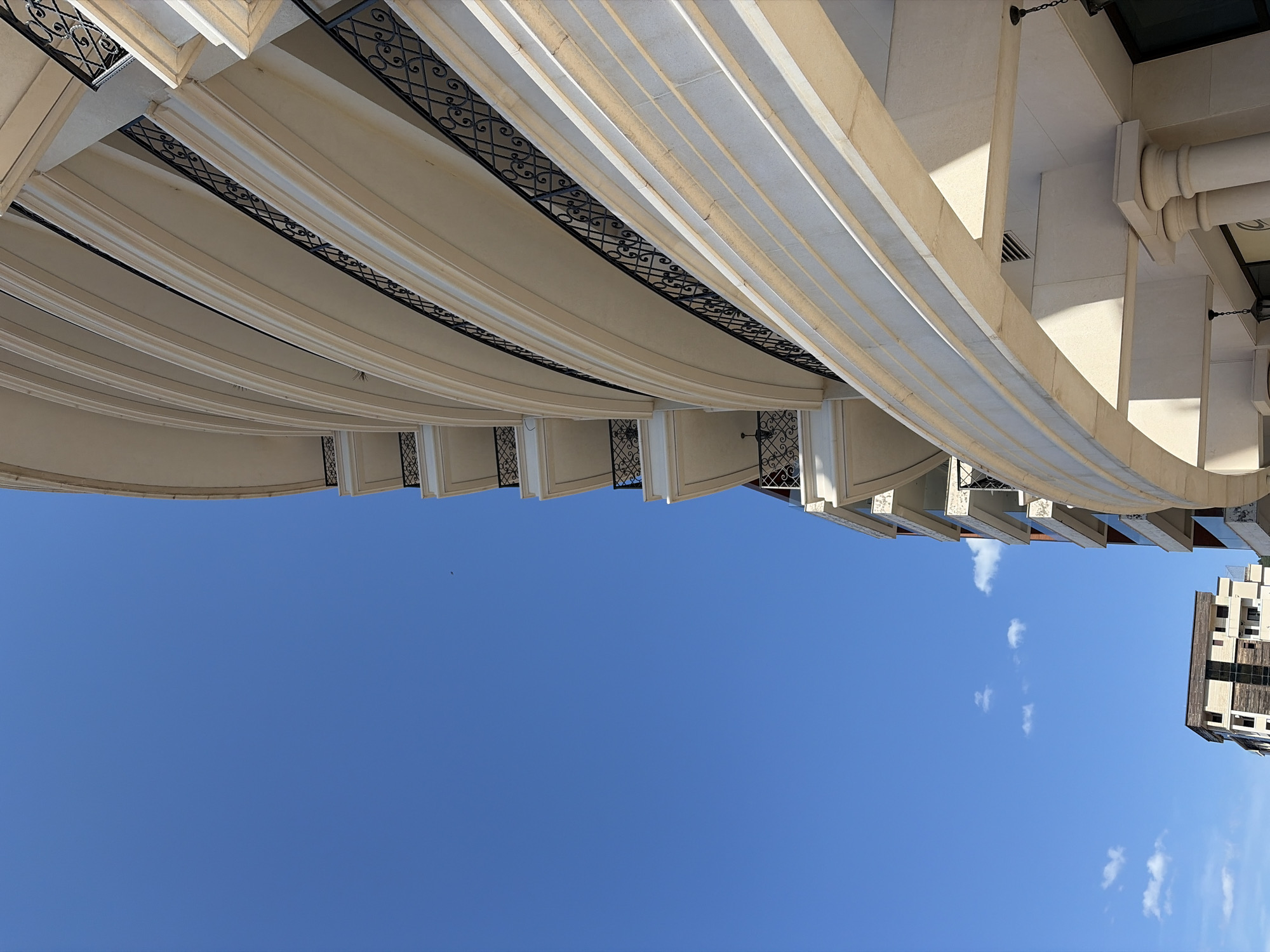 The image captures a view upwards towards the sky, framed by architectural elements. The main subject is a section of a building with a curved facade, featuring a series of balconies with decorative wrought-iron railings. The building appears to be constructed from light-colored stone, possibly limestone or a similar material, with contrasting dark metal railings. The sky is a clear blue with a few scattered white clouds. A partial view of another building can be seen in the upper right, revealing windows and a simple rectangular form. Given the style of architecture, it is highly likely that this image was taken in the city of Budva, Montenegro. The time of day appears to be daytime, and the light suggests clear weather conditions. The image is taken from a low angle.