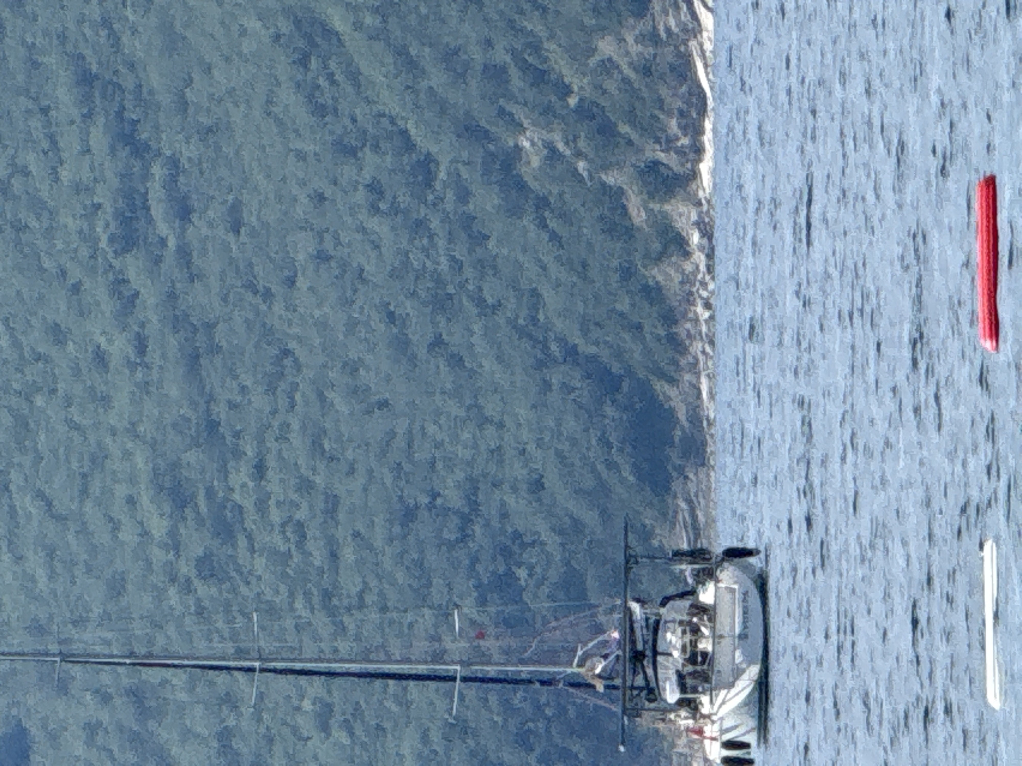 The image captures a sailboat on the Adriatic Sea, likely near Budva, Montenegro, given the coastal environment. The sailboat is white, and the mast has a visible black band. The foreground consists of the blue sea with a red buoy marking a channel. A strip of shoreline is visible, transitioning into green forested mountains, likely marking the end of the coastal area near Budva. The water surface has a slightly choppy texture.