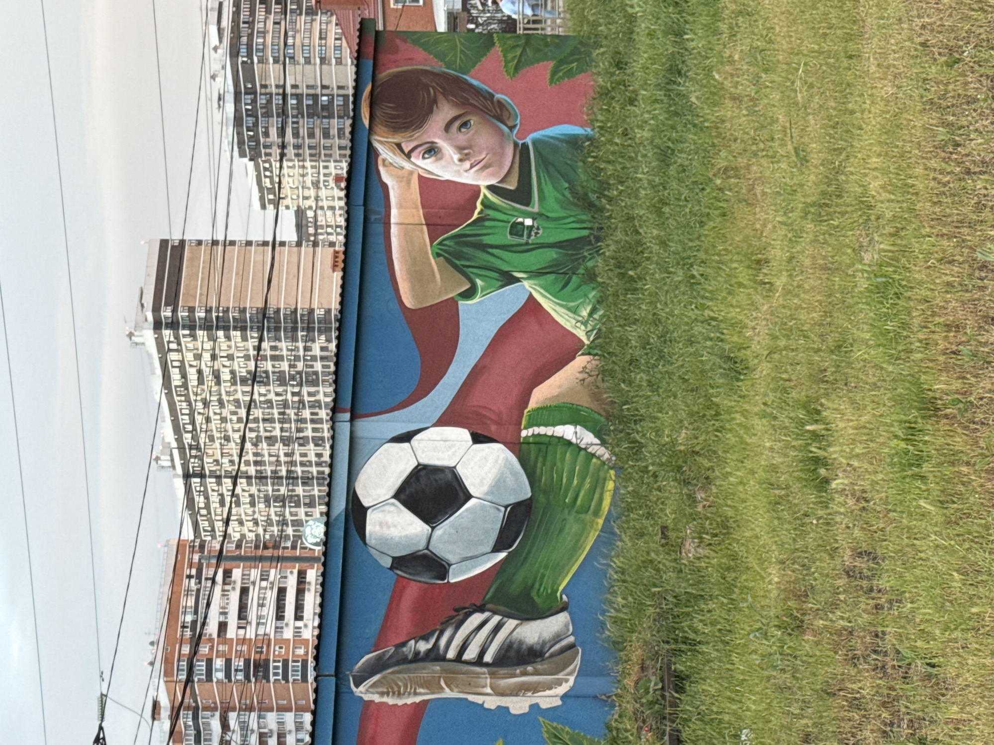 A mural is visible on the side of a building, portraying a young boy dressed in a green soccer jersey and socks, posing with a soccer ball. He has one hand resting near his head and is wearing a soccer shoe. Behind the boy is a background with red and blue colors. Adjacent to the mural is a patch of green grass. In the background, there are buildings resembling apartment complexes, and overhead power lines. The mural is located in the city of Krasnodar, Russia.