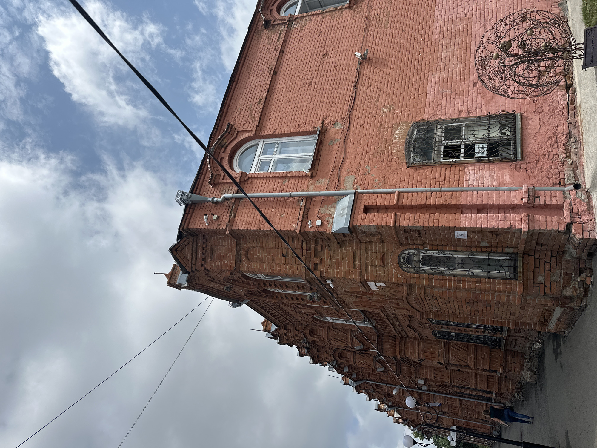 In Nogornyi, Russia, an old red brick building with ornate architectural details lines a street. The building features arched windows, some with decorative metal grates, and elaborate brickwork along its facade. A person is walking away from the camera down the sidewalk next to the building. The sky above is partly cloudy with a mix of blue sky and gray clouds. Power lines are visible stretched across the sky. A circular metal sculpture with a fruit-like design sits in front of the building. Security cameras are mounted on the building's exterior.
