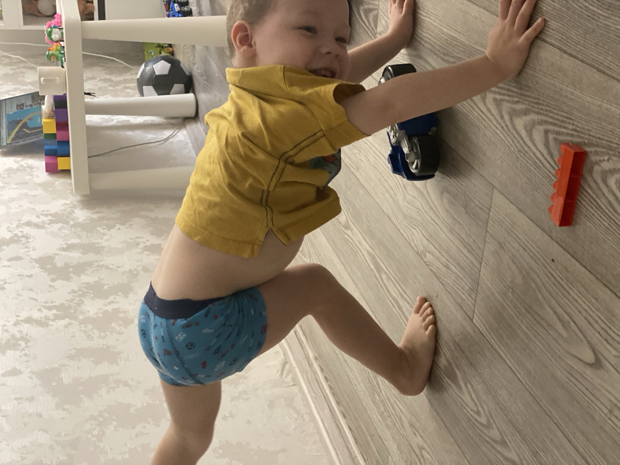 A child is seen indoors against a light-colored, wood-grain wall. The child is fair-skinned, wearing a mustard-colored shirt and blue underpants with a print. The child is positioned close to the wall, with one arm extended and a toy motorcycle adhering to the wall at approximately shoulder height. The child’s other arm is also raised to press against the wall. The child is standing with one foot against the wall, as if attempting to climb. To the right of the motorcycle is a red block toy. To the left, there's a shelving unit containing children's toys and a soccer ball. The location isn't deducible from the image.