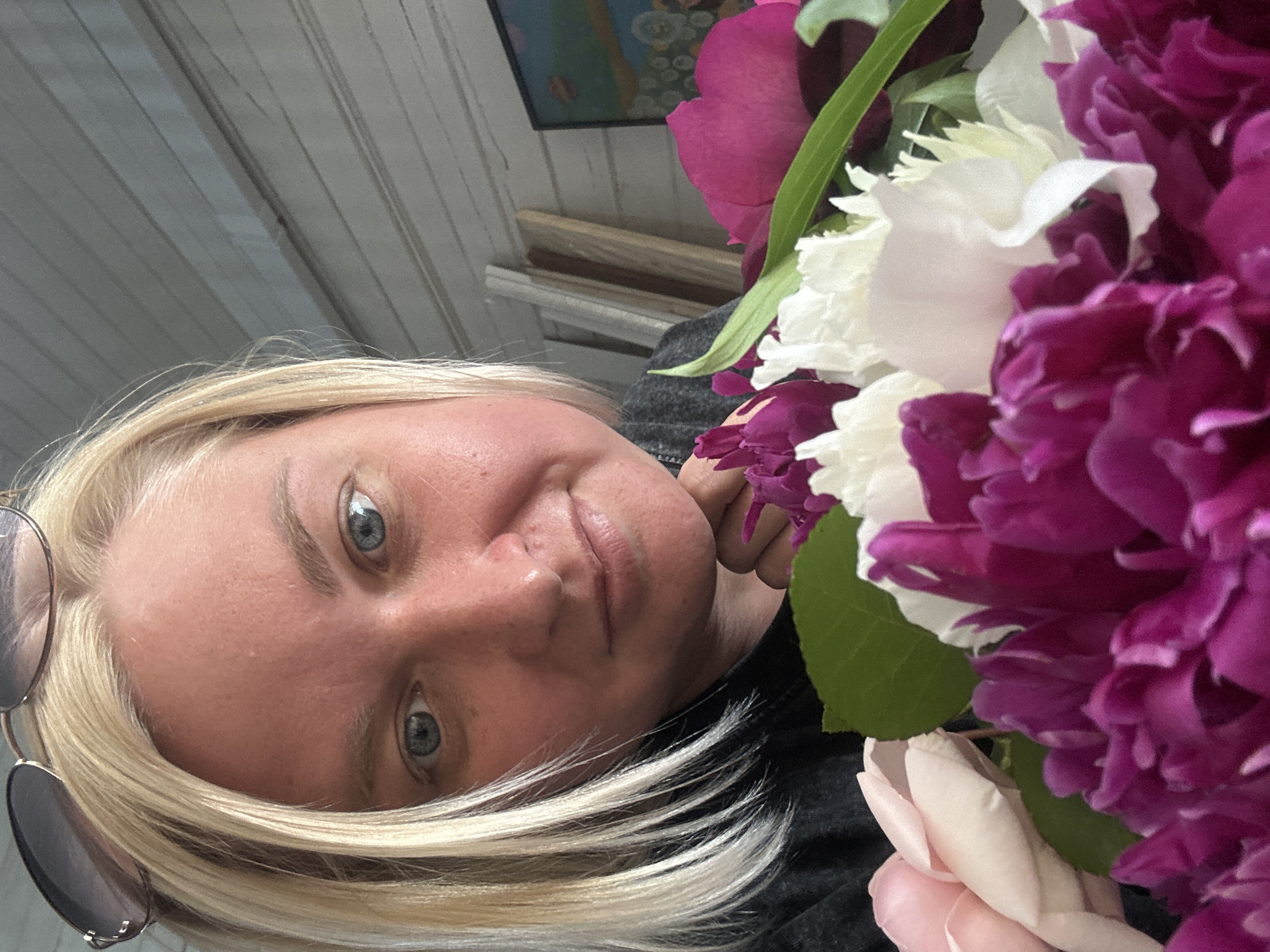 A close-up, rotated portrait shows a woman with blonde hair and blue eyes, holding a bouquet of flowers. The flowers are a mix of vibrant purple, white, and soft pink peonies and roses. She is wearing sunglasses on the left side of her head. Her expression is relaxed and natural, with a slight smile. The background appears to be a wooden structure, possibly a porch or an outdoor seating area, with white siding and a hint of a painting on the wall. The lighting suggests it might be daytime. The image is likely taken outdoors in a casual setting. There is no visible text.