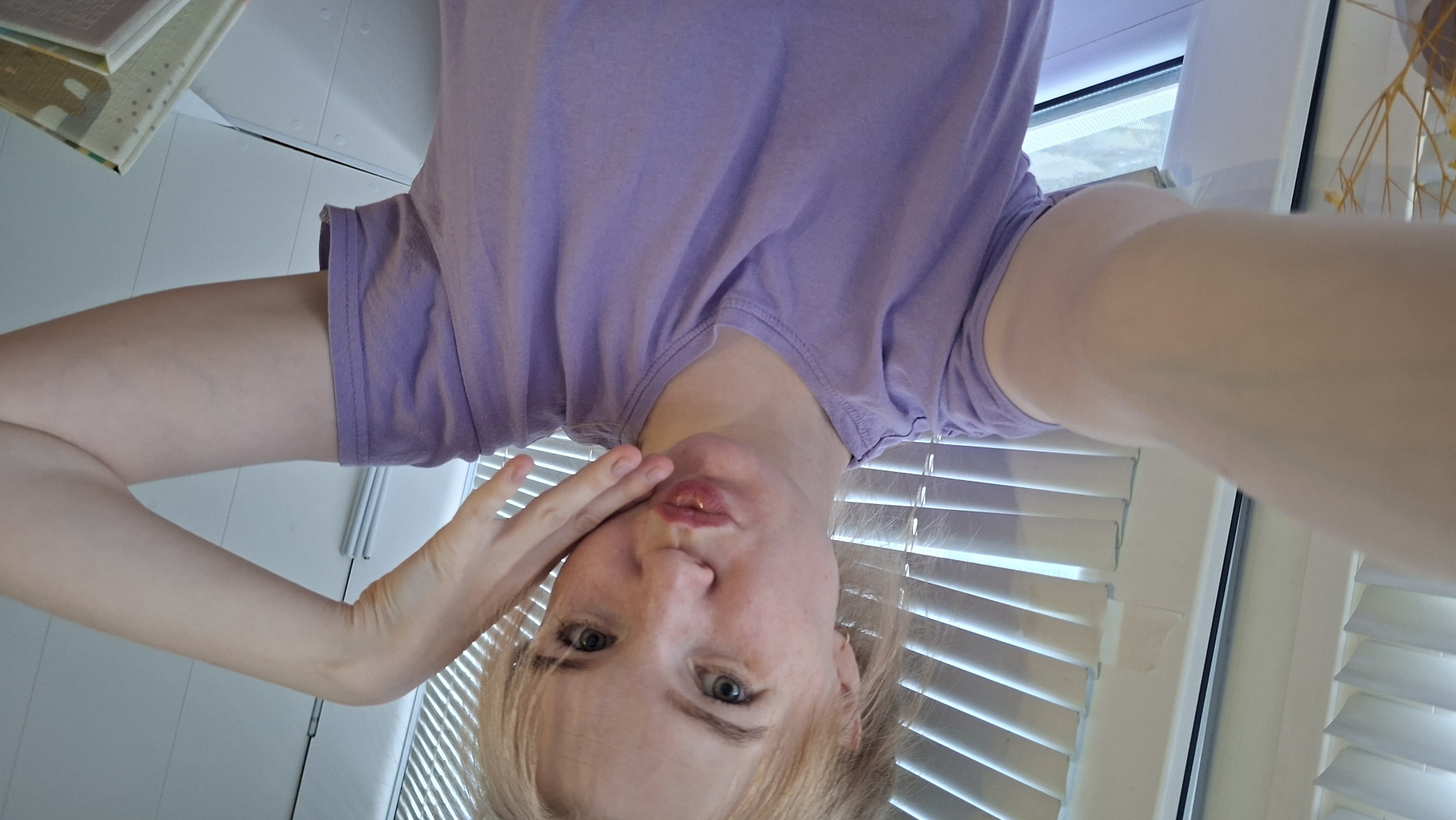 The image is a well-lit, close-up, self-portrait of a young, light-skinned female with light blonde hair, captured indoors. She has light-colored eyes and a fair complexion with visible freckles. Her hair, styled with a fringe, appears to be pulled back or short. She is wearing a plain lavender short-sleeved t-shirt with a rounded neckline. Her lips are pursed in a pouting expression, and her right hand rests gently against her cheek, fingers slightly spread. Her left arm is extended towards the camera, indicating she is holding the device herself.
In the background, horizontal white window blinds are visible, allowing diffused light into the interior space. A white window frame is partially visible on the left side of the frame, and a light-colored wall or interior surface is seen on the right. In the lower right corner, the corner of what appears to be a stack of books or notebooks with patterned covers is visible. On the far left edge, a few dry, thin plant stems or decorative elements are partially visible. The specific geographical location, including the city of Chaikovsky, Russia, cannot be determined from the visual information provided within the image.