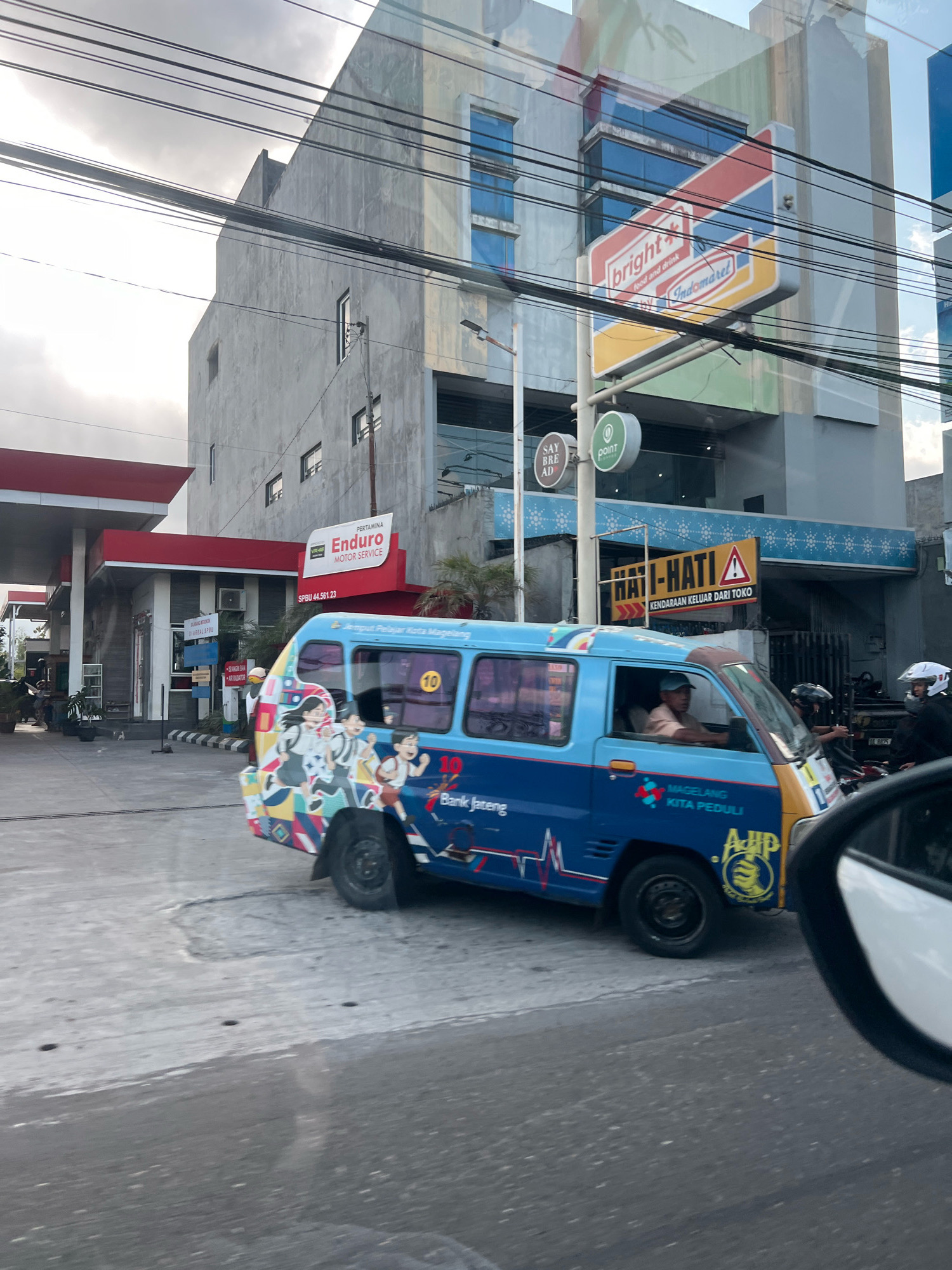 The image depicts a street scene in Magelang, Indonesia, under an overcast sky. In the foreground, a blue and yellow van with anime-style graphics on its side is parked. The van has the text "Bank Jateng" and "MAGELANG KITA PEDULI" written on it, indicating its affiliation with a bank and a local initiative in Magelang. A man in a light-colored shirt and blue cap is visible driving the van. Several motorcycles are also present, some with riders wearing helmets.
Behind the van, a gas station with red and white awnings and a "PERTAMINA ENDURO MOTOR SERVICE" sign is visible. Adjacent to the gas station is a multi-story building with a prominent "Indomaret" and "bright" convenience store sign. Another sign reads "HATI-HATI" (Be Careful) with an exclamation mark and the text "KENDARAAN KELUAR DARI TOKO" (Vehicles exiting the store). The environment appears to be a commercial area with buildings and overhead power lines, typical of a city in Indonesia. The overall impression is that of a busy urban street with commercial establishments.