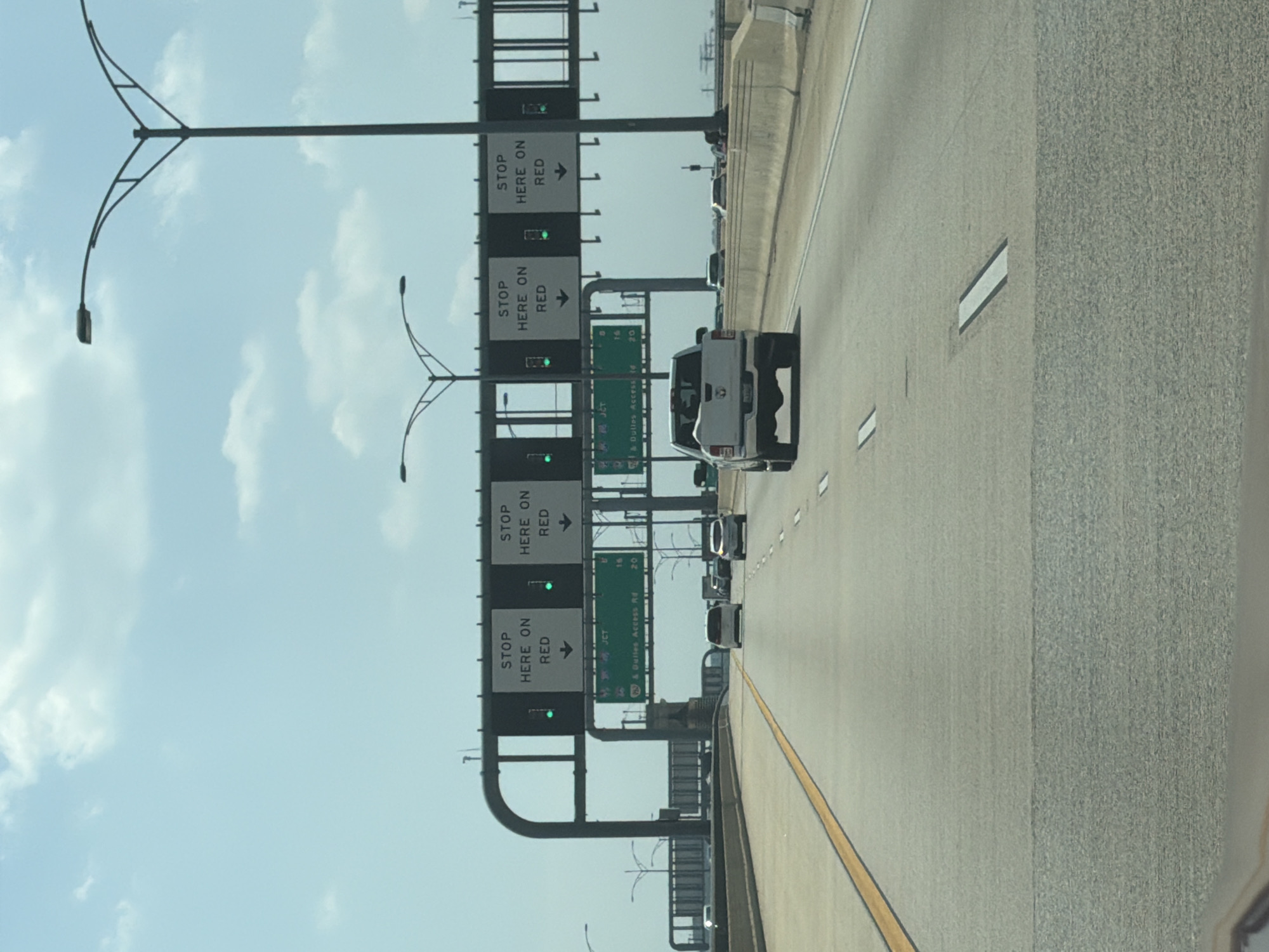 An eye-level, medium shot captures a view of traffic moving on a highway in Oxon Hill, Maryland. Overhead signs display "STOP HERE ON RED" in white lettering against black backgrounds, with downward-pointing arrows. Green traffic lights are illuminated below these signs. A white pickup truck is the closest vehicle, followed by a white SUV and a dark-colored SUV. The road is paved, with a yellow line marking the left lane and white dashed lines separating lanes. The sky is a clear blue with scattered white clouds, and streetlights are visible on both sides of the highway.