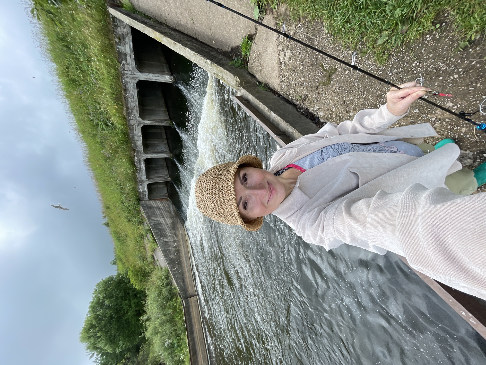 In the city of Bereznik, Russia, a woman is taking a selfie by a water canal. She is wearing a light-colored button-up shirt over a pink and gray top, a straw hat, and green boots. She is holding a fishing rod, and it appears she is fishing. The water in the canal is flowing rapidly, and there is a series of concrete culverts in the background. The sky is overcast, and there is a bird flying in the distance. The area around the canal is covered in grass and some concrete pathways.
