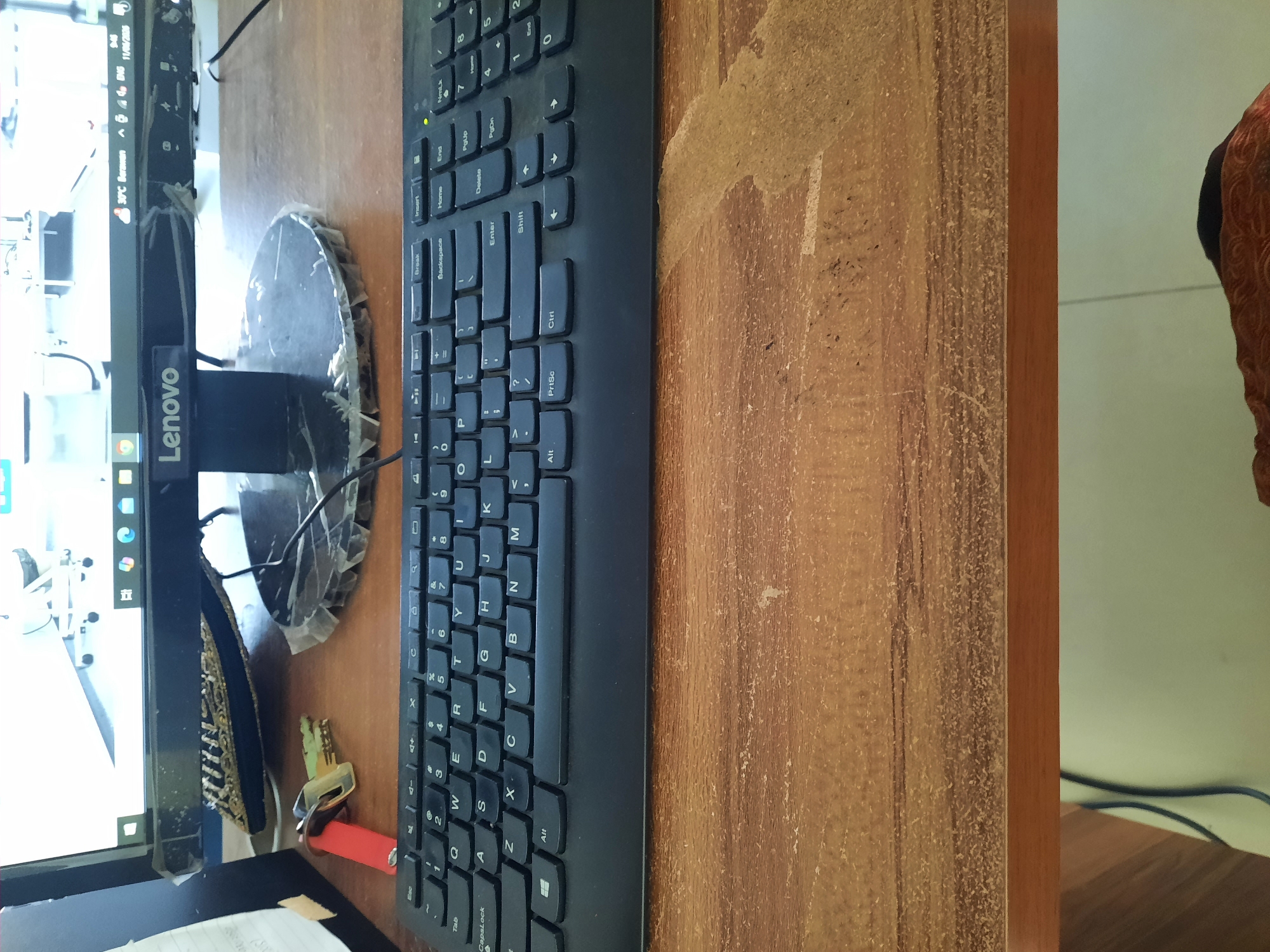 The image captures a close-up, slightly angled view of a desk with a Lenovo monitor and a black keyboard. The monitor displays a screenshot of a web browser with several tabs open, including what appears to be a simulation of airport vehicles on a runway. The bottom of the monitor shows the time as 9:46 AM, the date as 11/06/2025, and a temperature of 30°C in Lhokseumawe, Indonesia.
On the desk, to the left of the keyboard, are a red lighter and two keys. The keyboard itself is a standard QWERTY layout. The desk surface is wooden with a worn, textured finish, showing signs of use and some light debris. The overall impression is of a functional, lived-in workspace. There are no people visible in the image. The context of Lhokseumawe, Indonesia, is provided by the monitor's display.