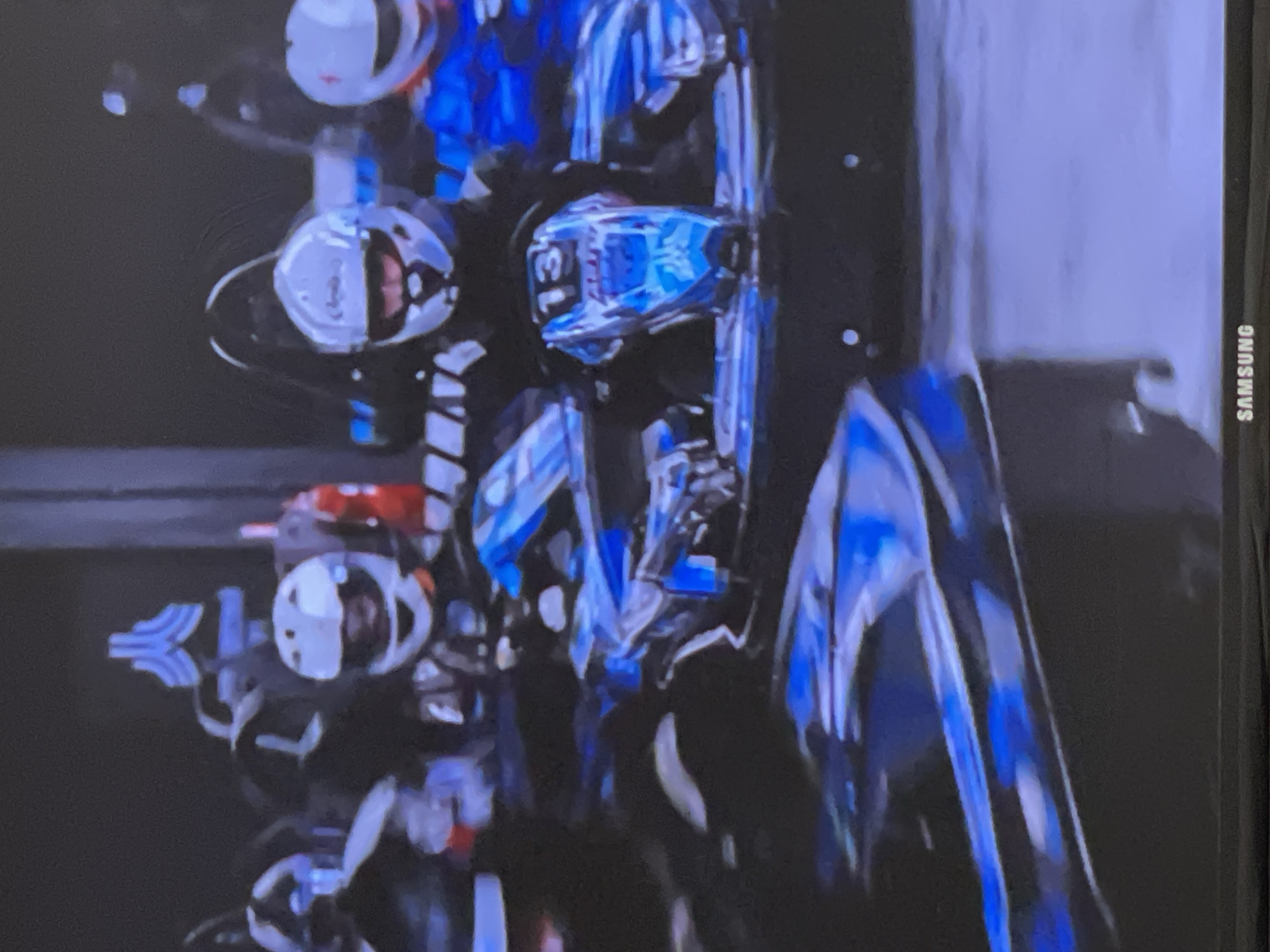 The image displays several individuals, likely racers or motorcycle enthusiasts, wearing full-face helmets and protective gear, alongside high-performance racing motorcycles or similar vehicles. The scene is set against a dark, undifferentiated background, suggesting an indoor event, a staging area, or a nighttime gathering in Moscow, Russia.
In the foreground, a rider is visible wearing a white helmet with prominent blue and black accents. The number "13" is clearly displayed on the chin guard of this helmet, accompanied by other unidentifiable blue text or logos on the side. Another rider's white helmet with orange or red highlights is partially visible behind and to the left of the central figure. Further back, a third person is seen in a white helmet. The racing vehicles themselves are predominantly blue and black, featuring sleek, futuristic designs and reflective surfaces, with some white and red elements integrated into their bodywork. A distinctive stylized blue and white logo, resembling a "K" or a winged symbol, is visible on the left side of the frame, behind one of the riders.
The overall impression is that of an organized event, possibly a display, a pre-race lineup, or a post-race presentation of the riders and their machines. The image appears to be a photograph of a screen, evidenced by the "SAMSUNG" brand logo visible vertically along the right edge of the frame.